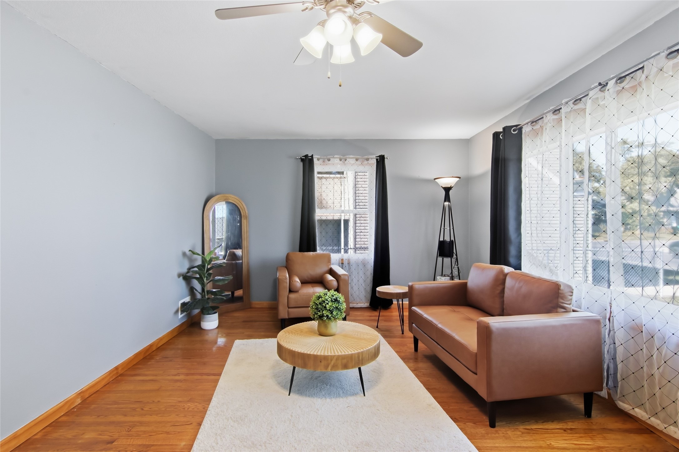 Living room with brown leather sofa and armchair, round wood coffee table, and large curtained windows.