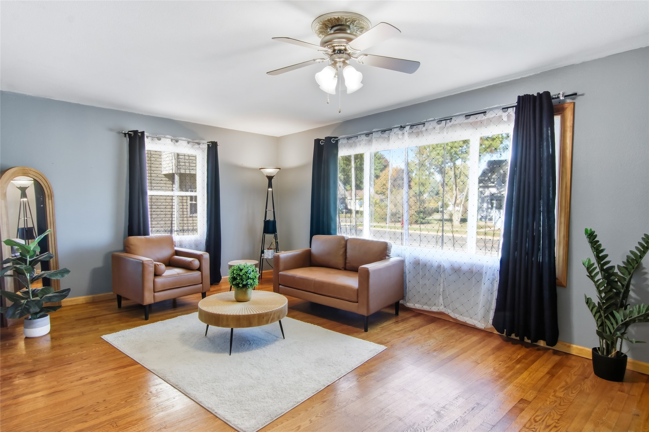 Living room with brown leather seating, hardwood floors, and a large curtained window.