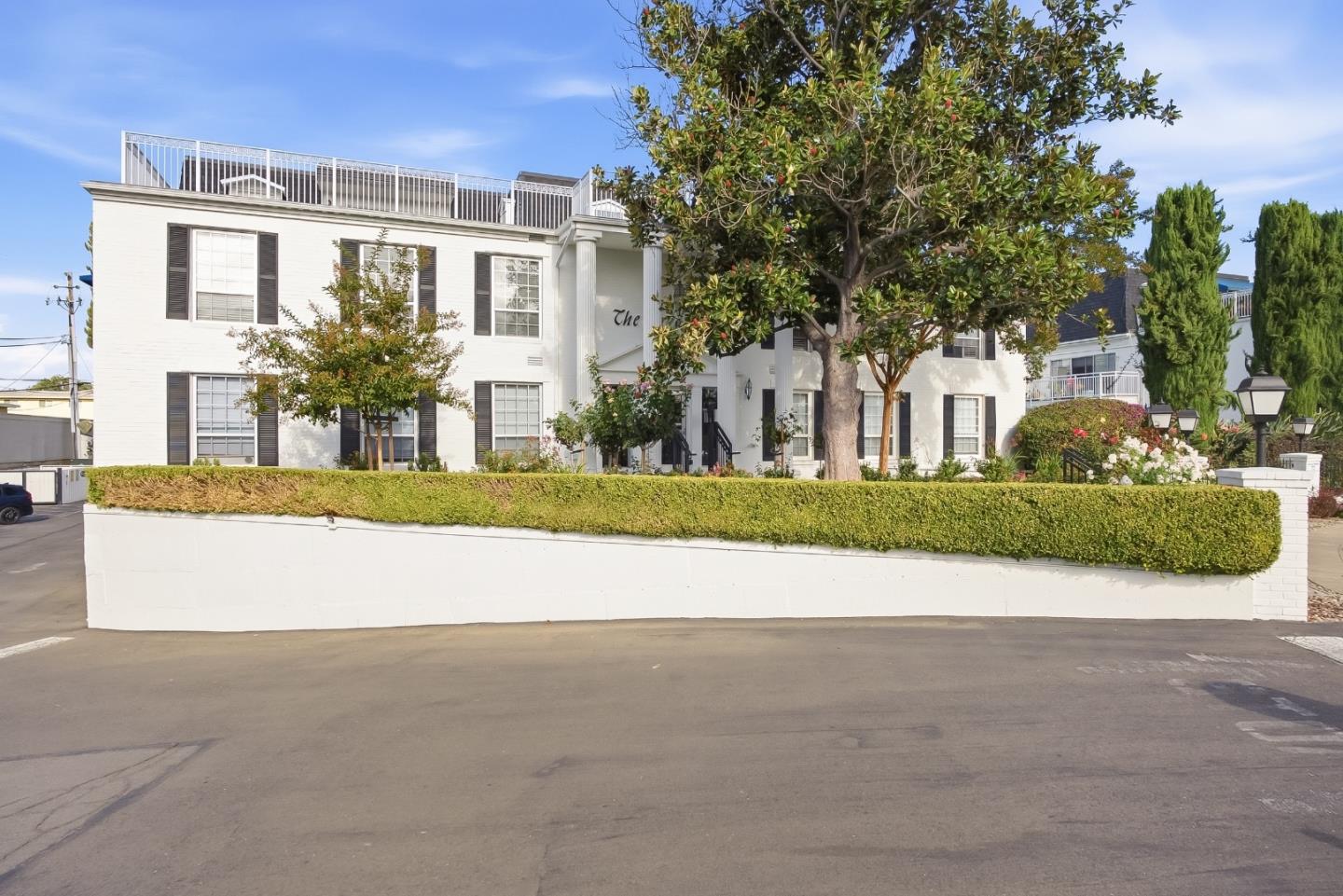 White multi-story residence with rooftop terrace, mature trees, and a clipped hedge along the street.