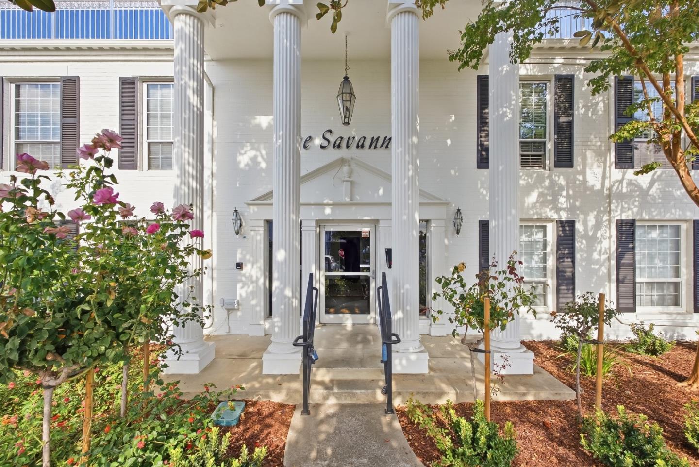 White residential building entrance with tall columns, a glass door, handrails, and landscaped flower beds.