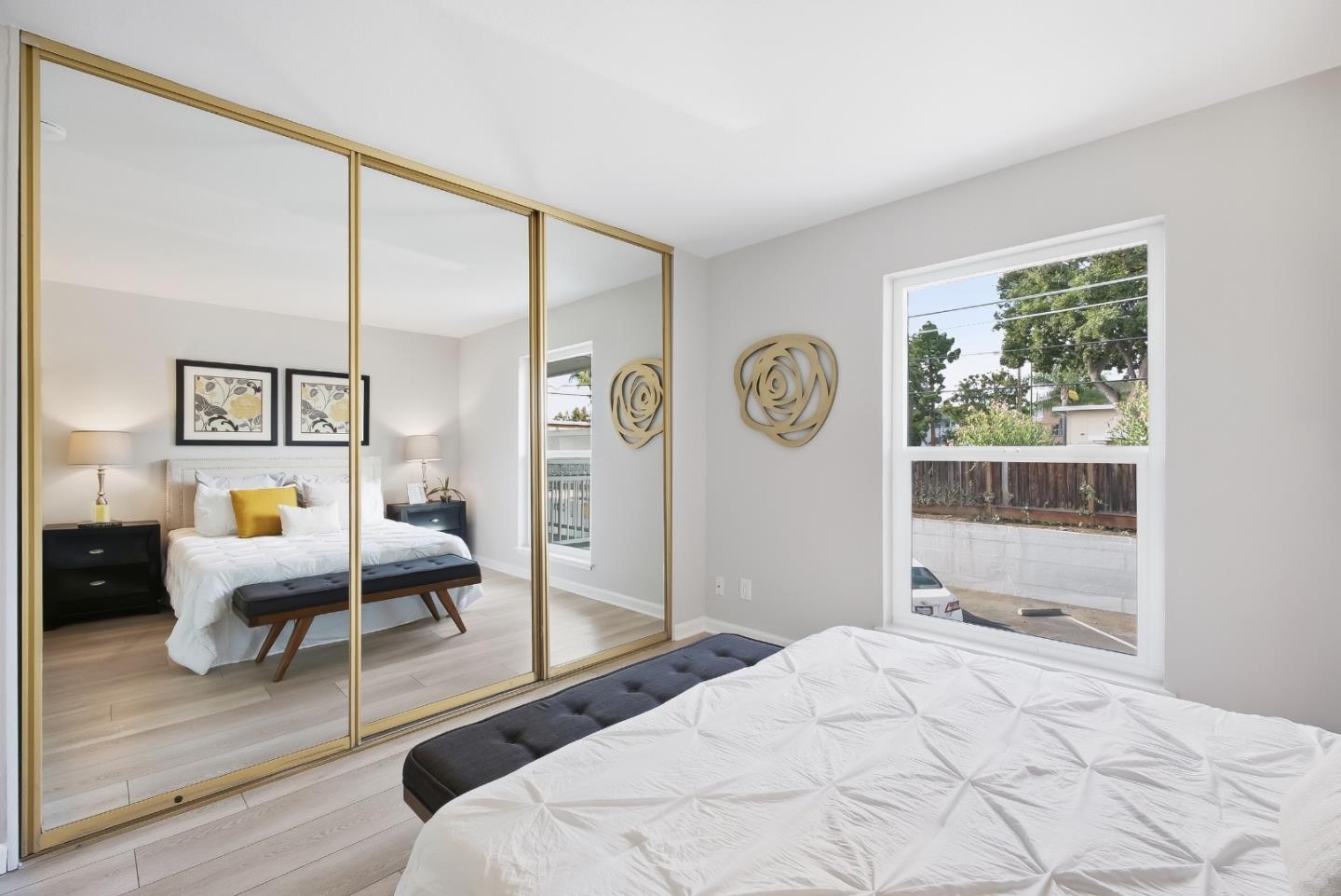 Staged bedroom with mirrored gold-framed closet doors, a white bed, and a large street-facing window.
