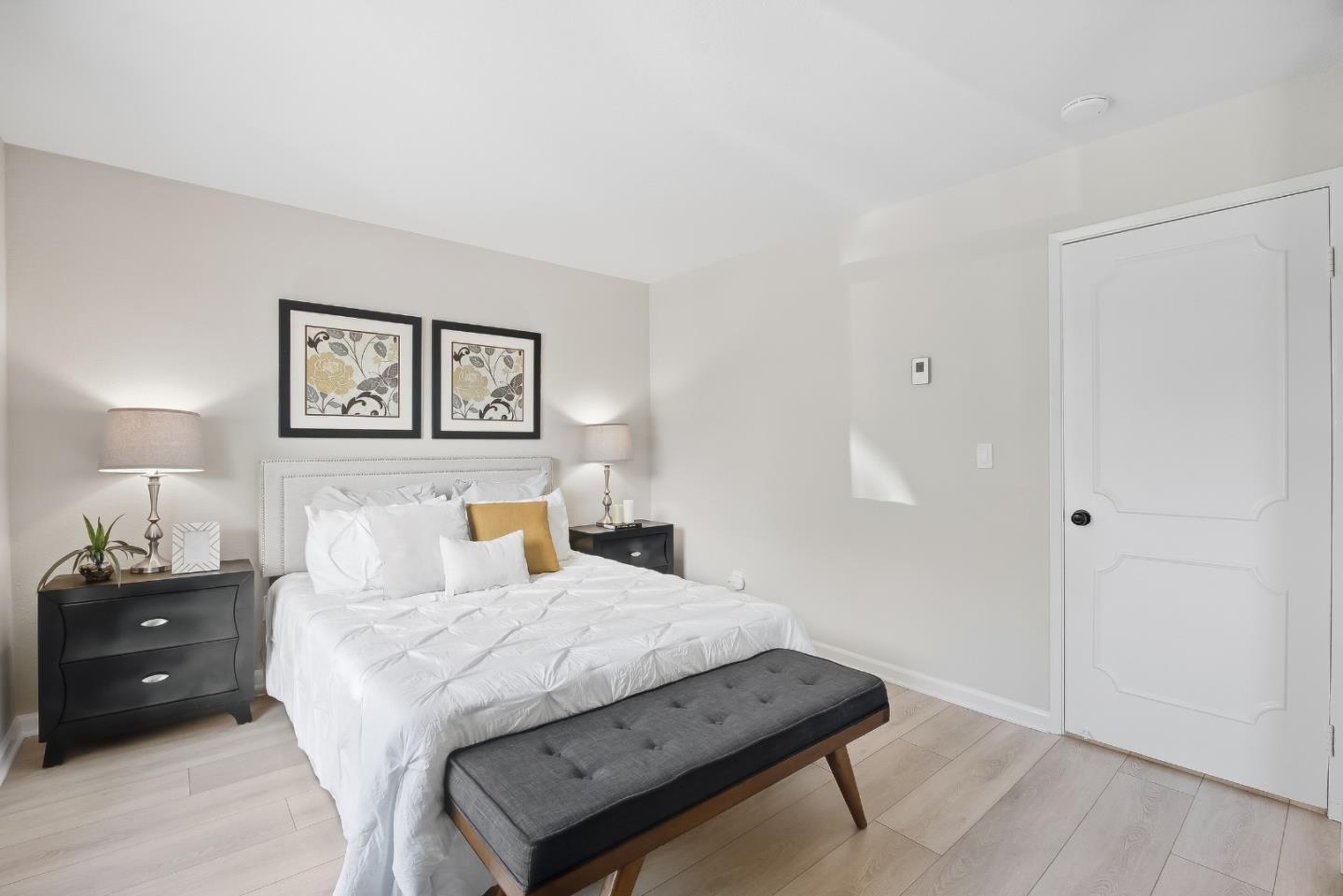 Staged bedroom with a white bed, floral prints, dark nightstands, and a charcoal bench.