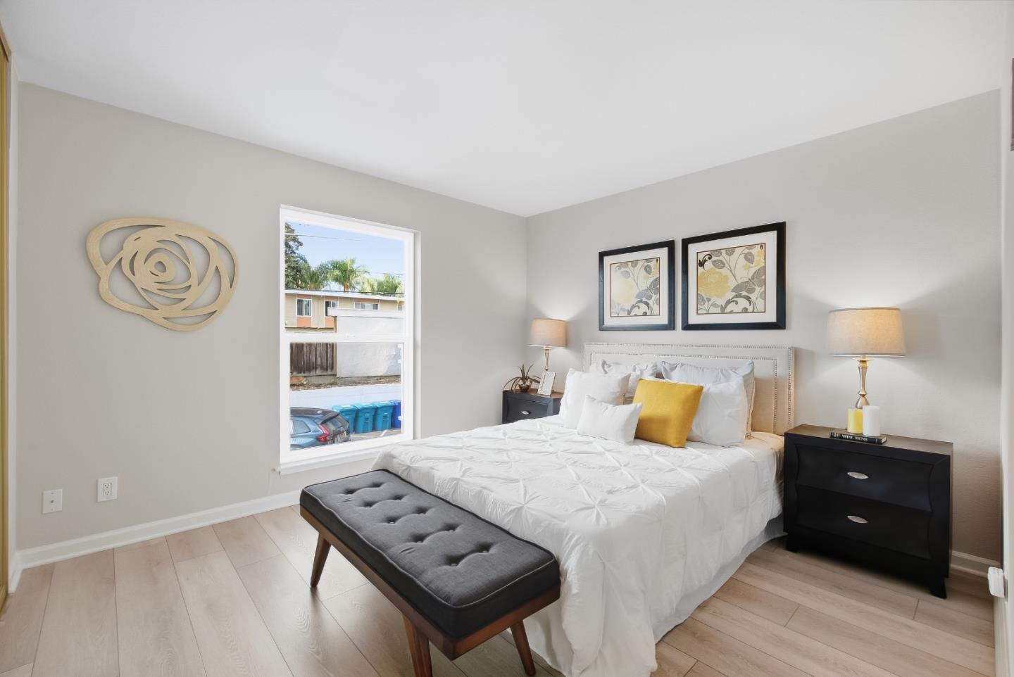 Staged bedroom with a white bed, gray bench, black nightstands, and a large window.
