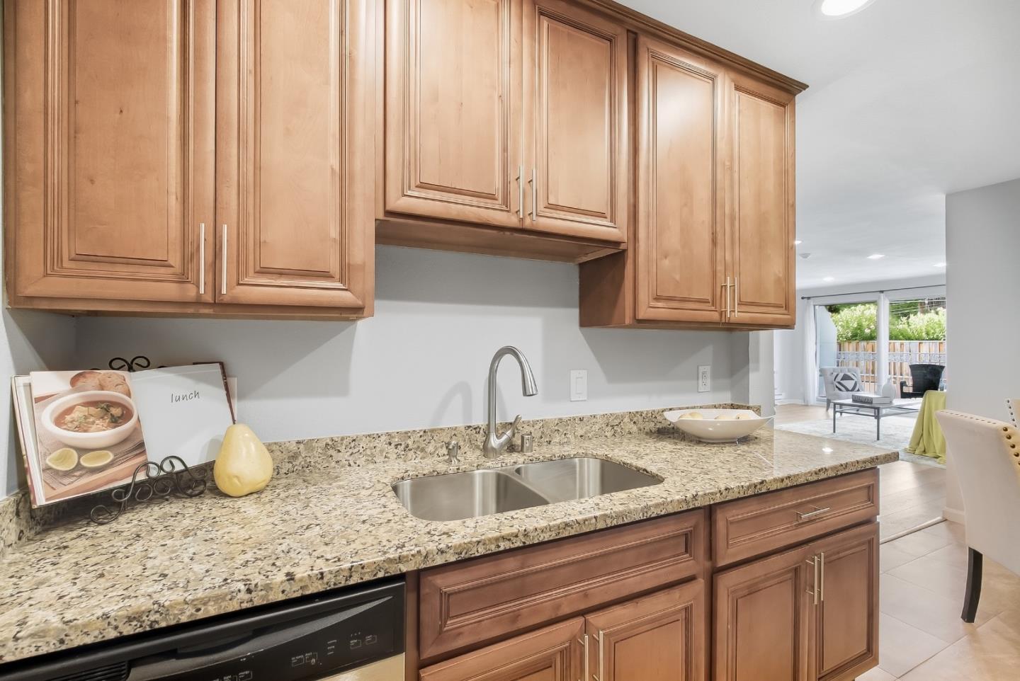 Kitchen with warm wood cabinets, granite countertops, and a double-basin sink opening to a bright living area.
