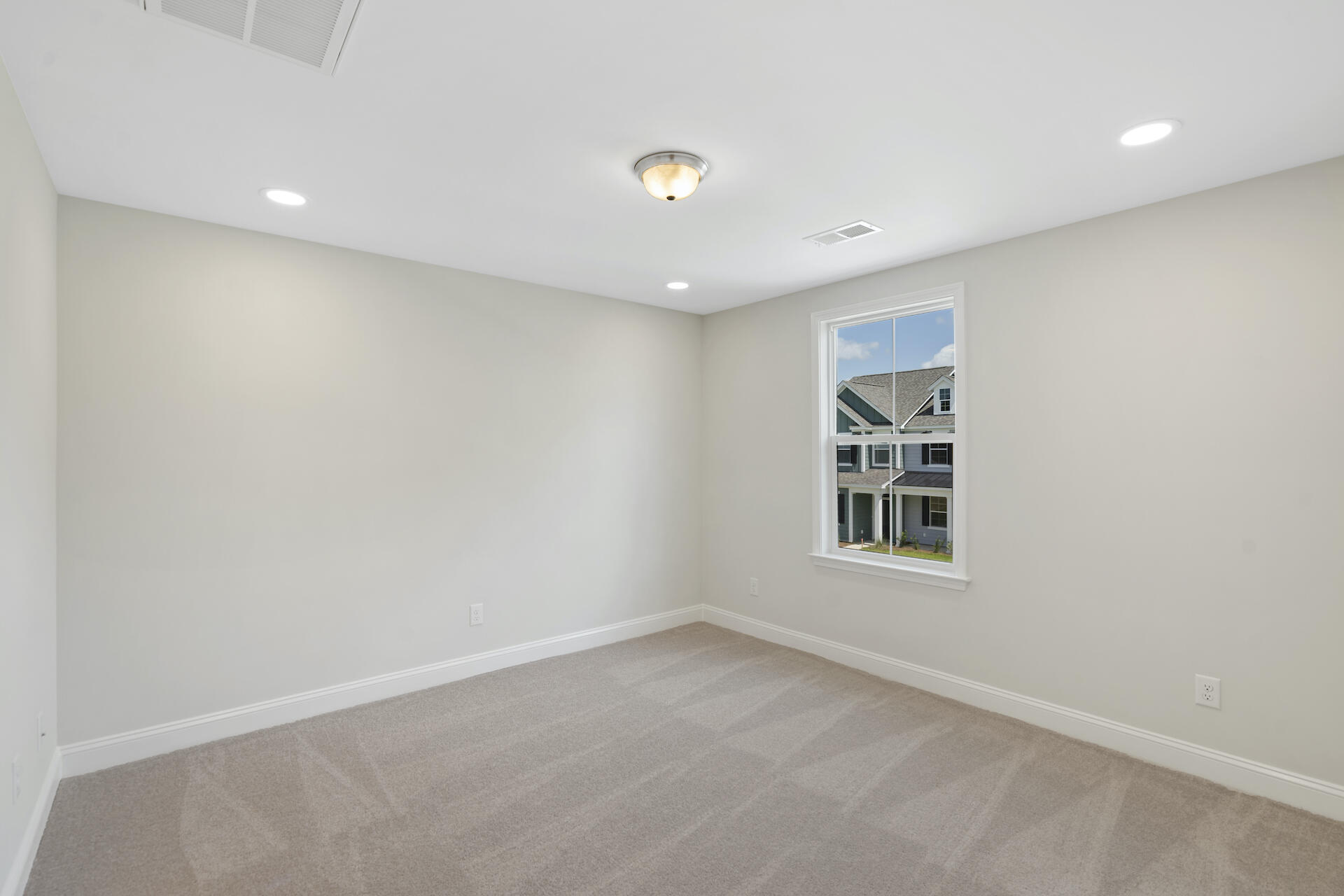 Empty carpeted room with neutral walls and a window overlooking neighboring houses.
