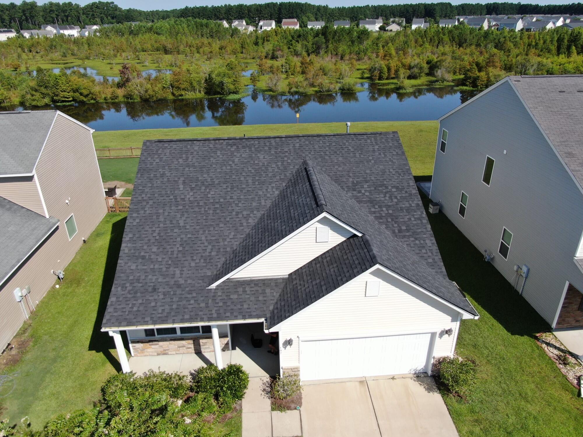 Aerial view of a house beside a pond, showing its dark roof, front porch, and two-car garage.