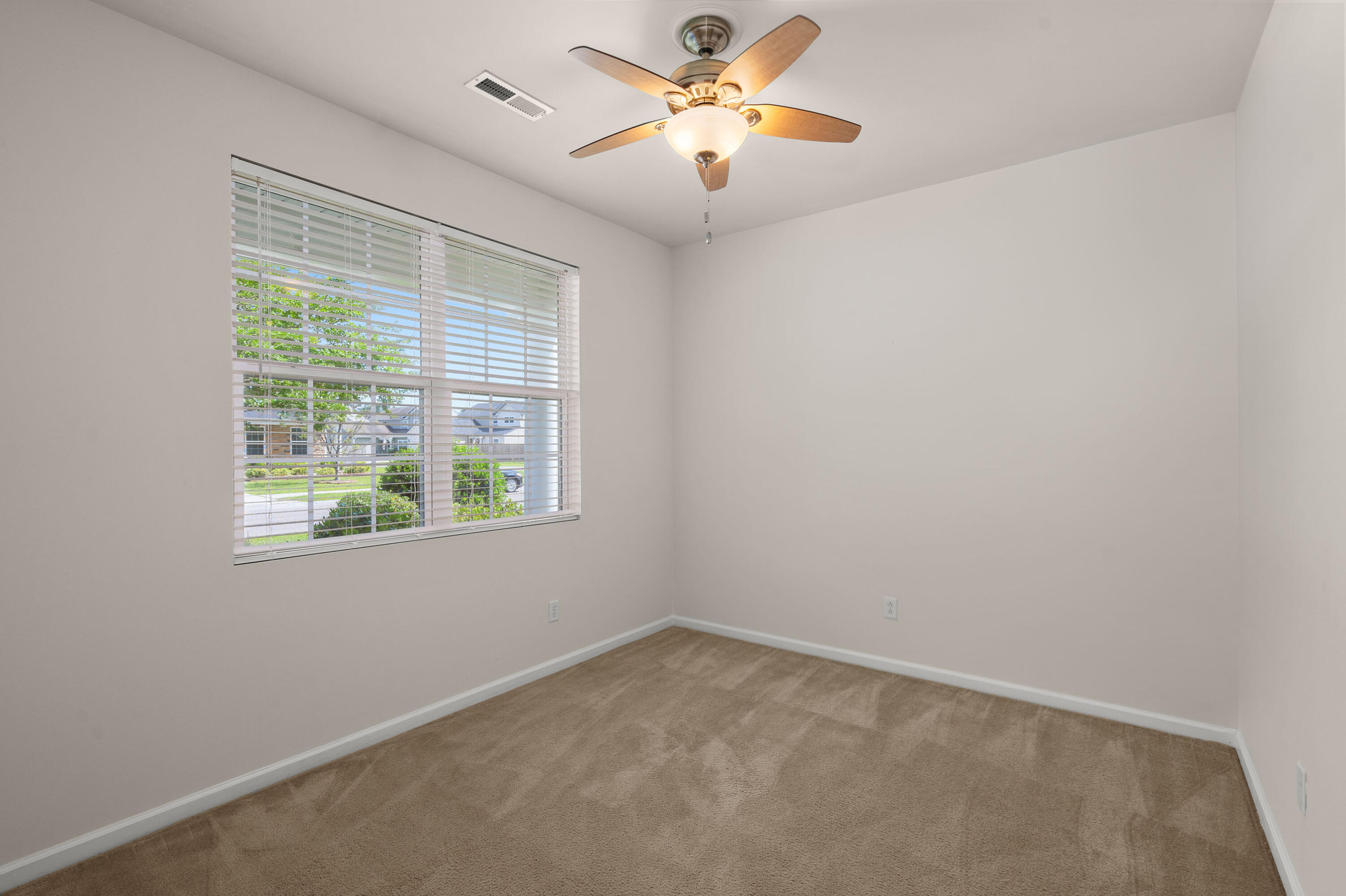 Empty carpeted room with a ceiling fan and a large window overlooking trees and neighboring homes.