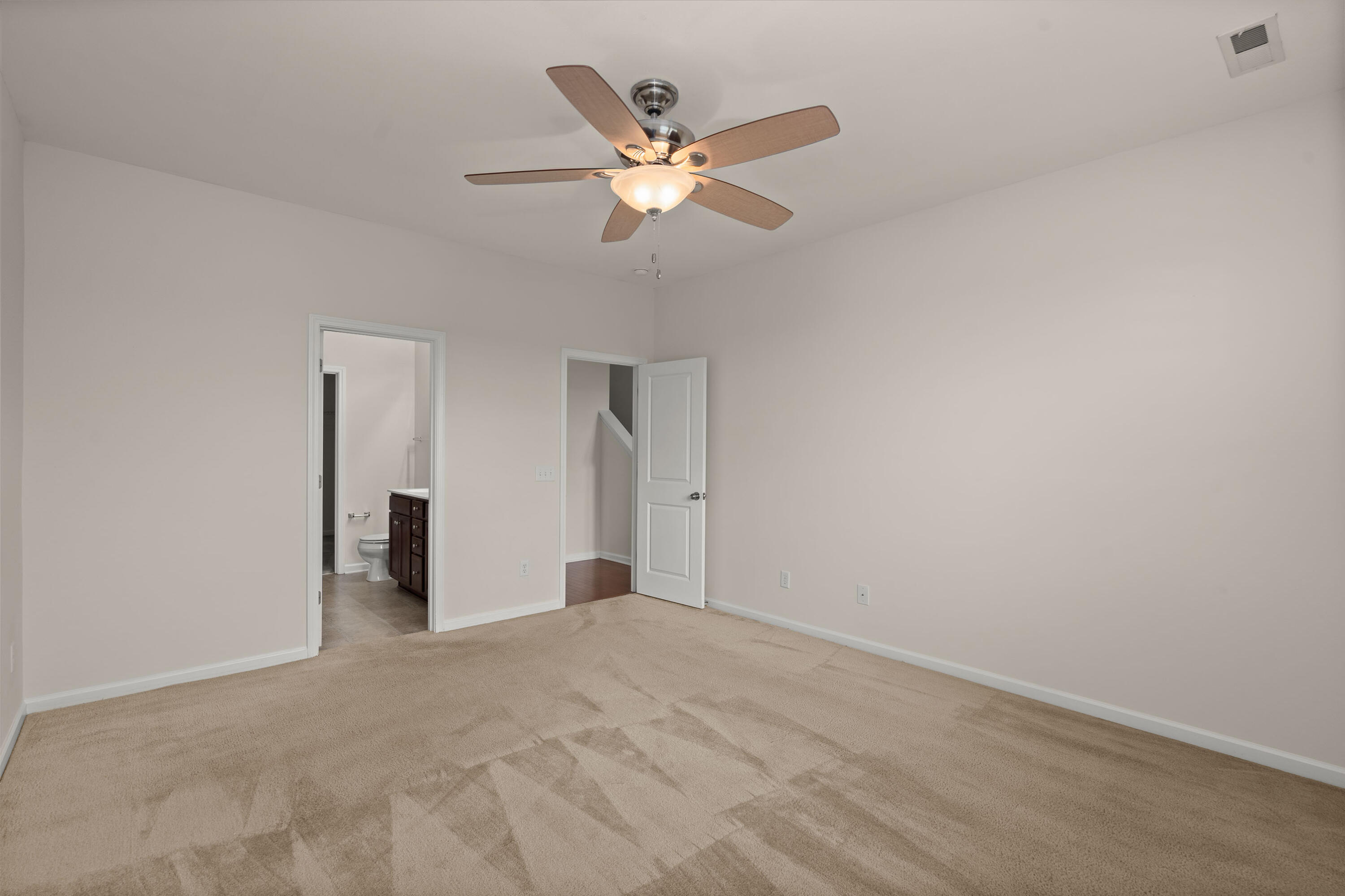 Empty carpeted bedroom with a ceiling fan and open doorways to a bathroom and stair hall.