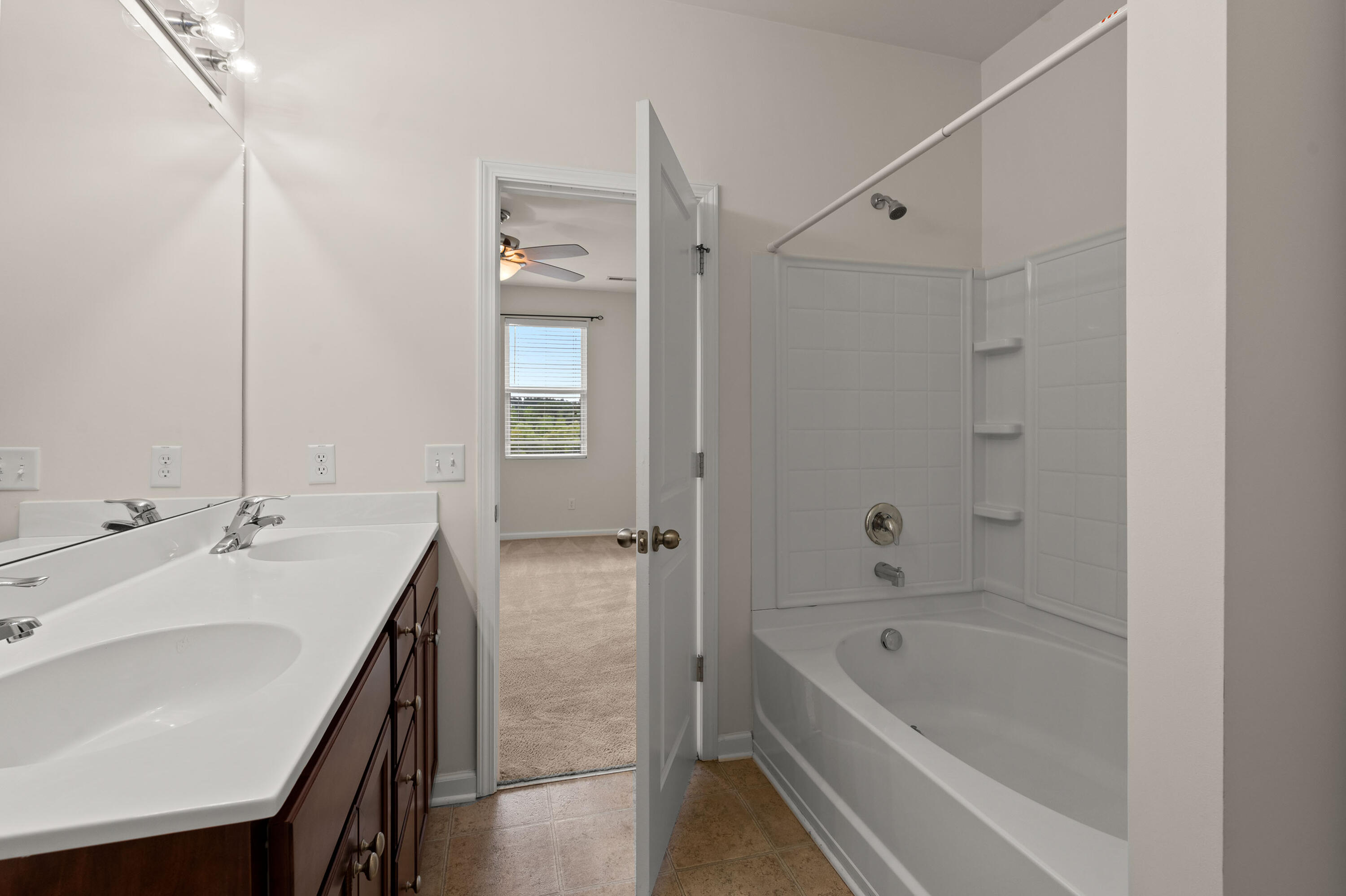 Bathroom with a double-sink vanity and white bathtub-shower beside an open doorway to a carpeted room.
