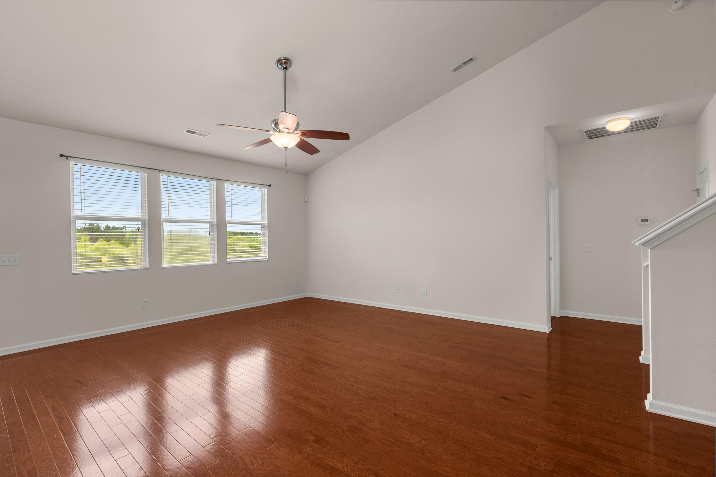 Empty living room with hardwood floors, three windows, a ceiling fan, and a staircase.
