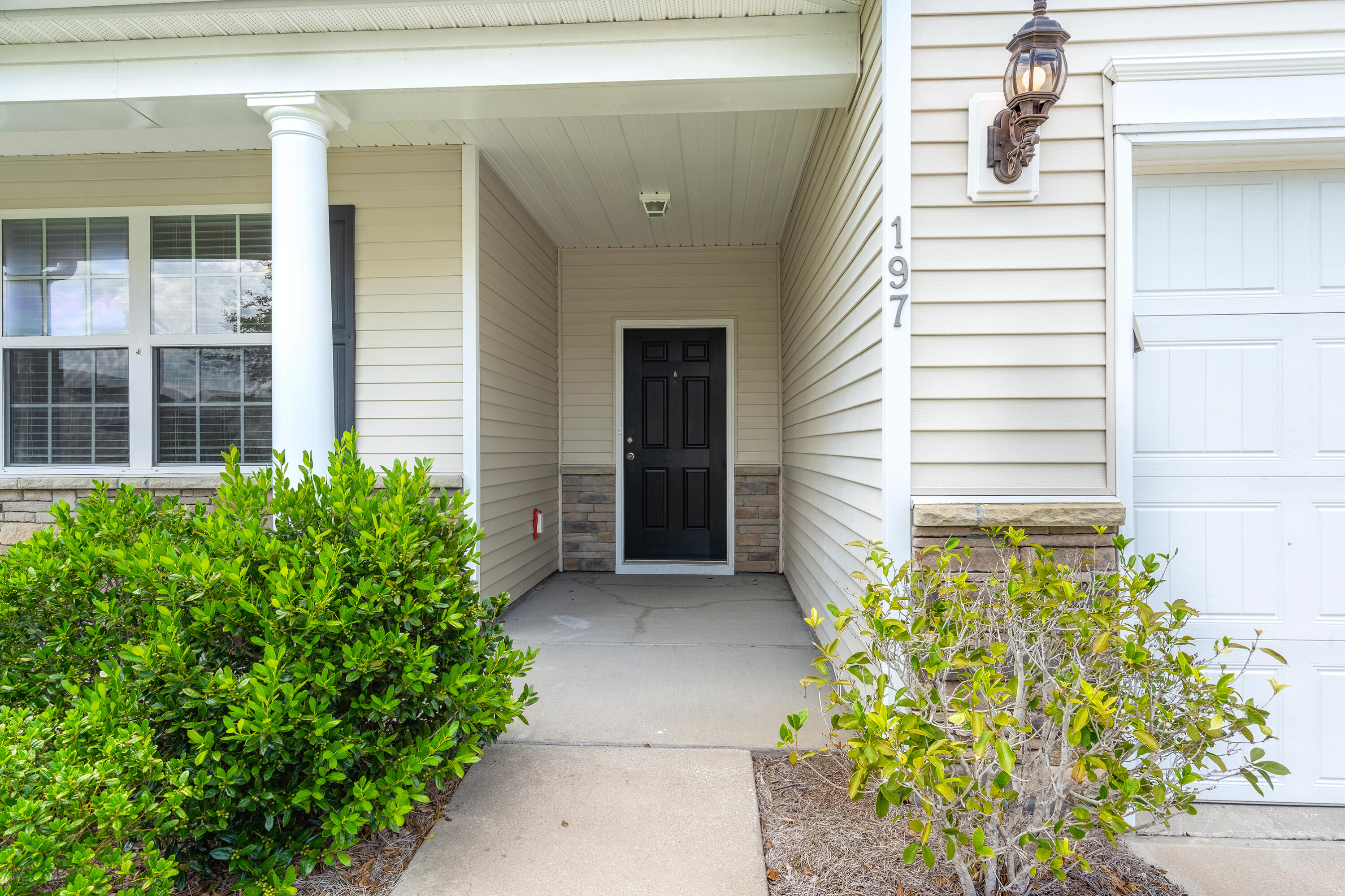 Covered front entry with a black door, white column, stone accents, and shrubs.