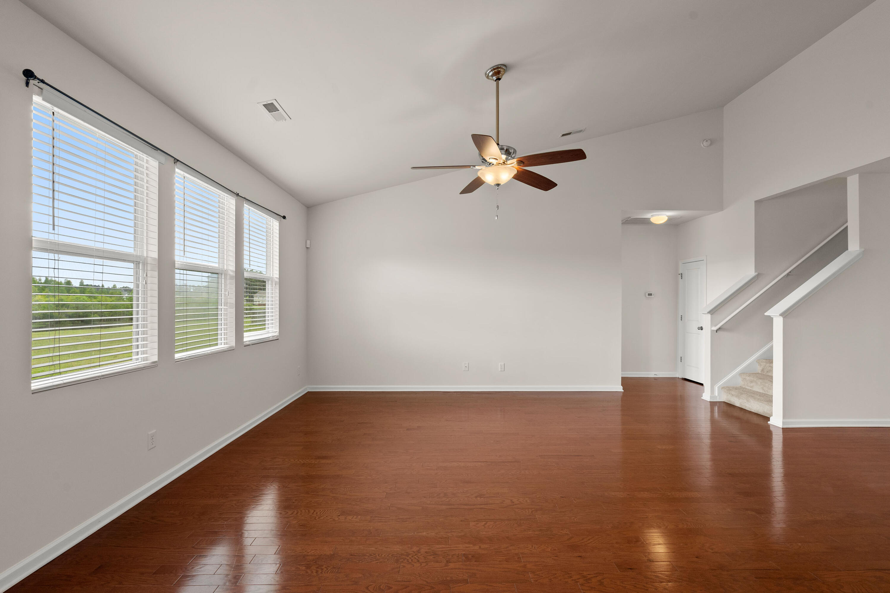 Empty living room with hardwood floors, three tall windows, a ceiling fan, and a staircase.