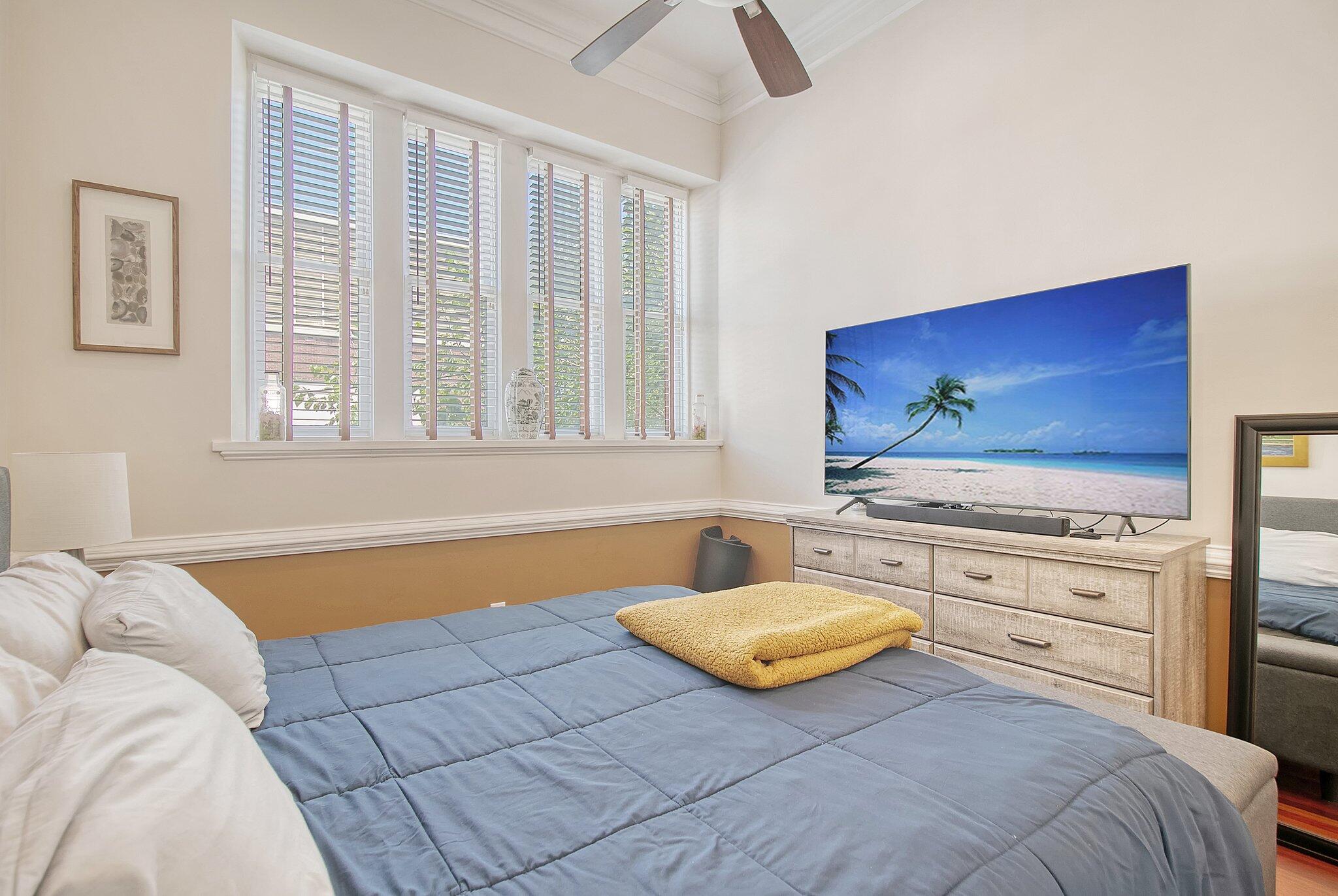 Bedroom with blue bedding, a folded yellow blanket, tall shuttered windows, and a television on a dresser.