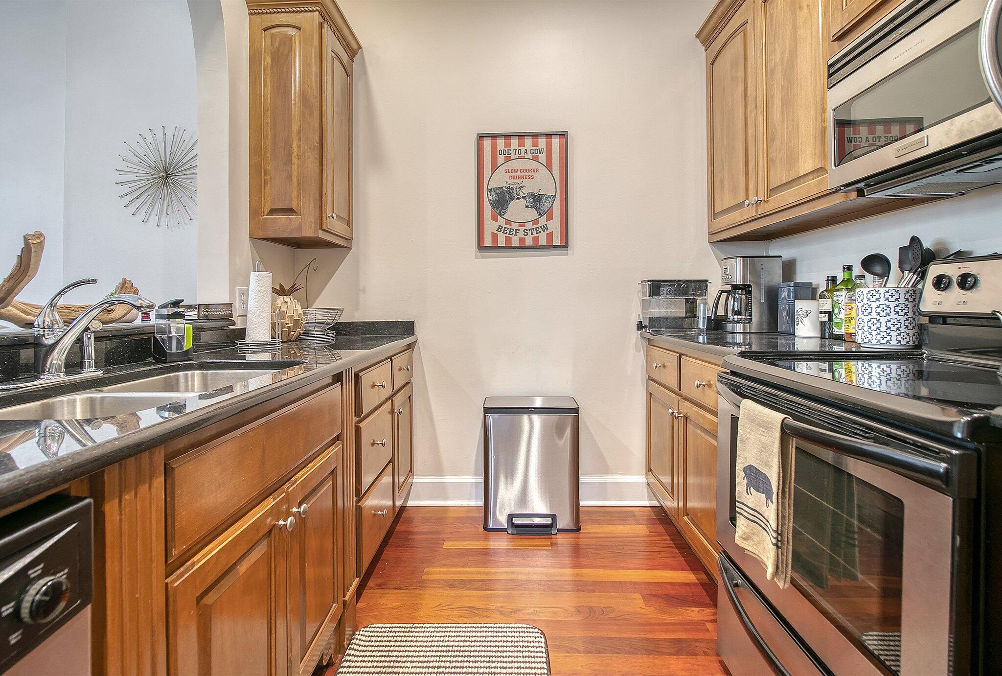 Galley kitchen with warm wood cabinets, black countertops, stainless steel appliances, and hardwood floors.