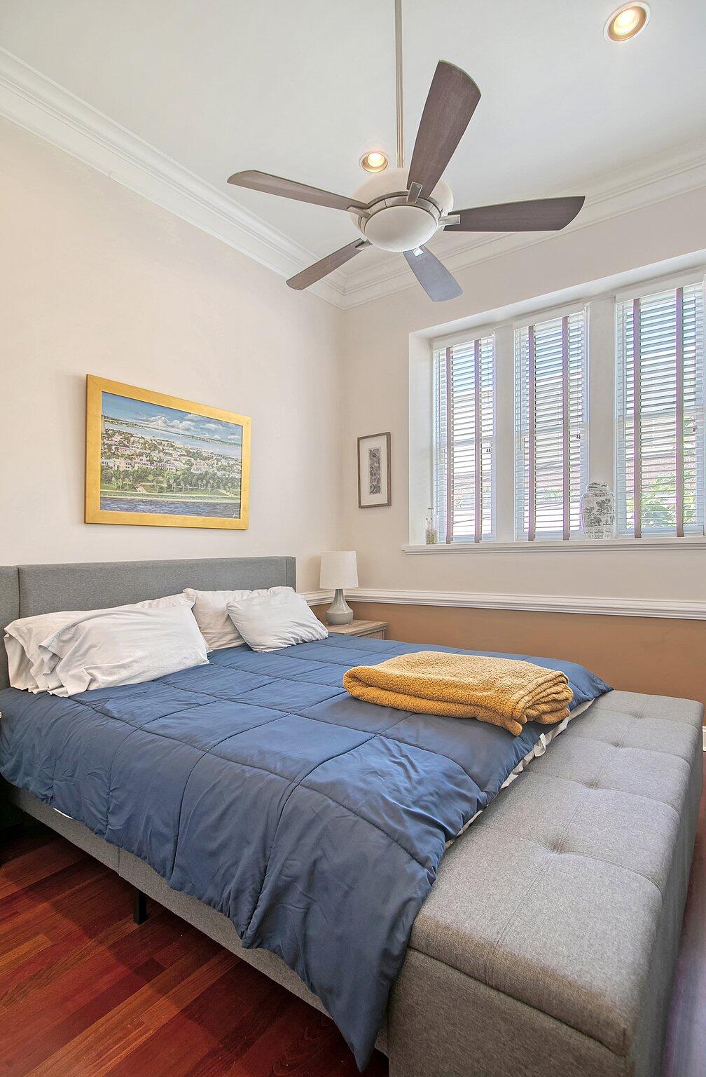 Bedroom with blue bedding, gray upholstered bed, plantation shutters, and a ceiling fan.