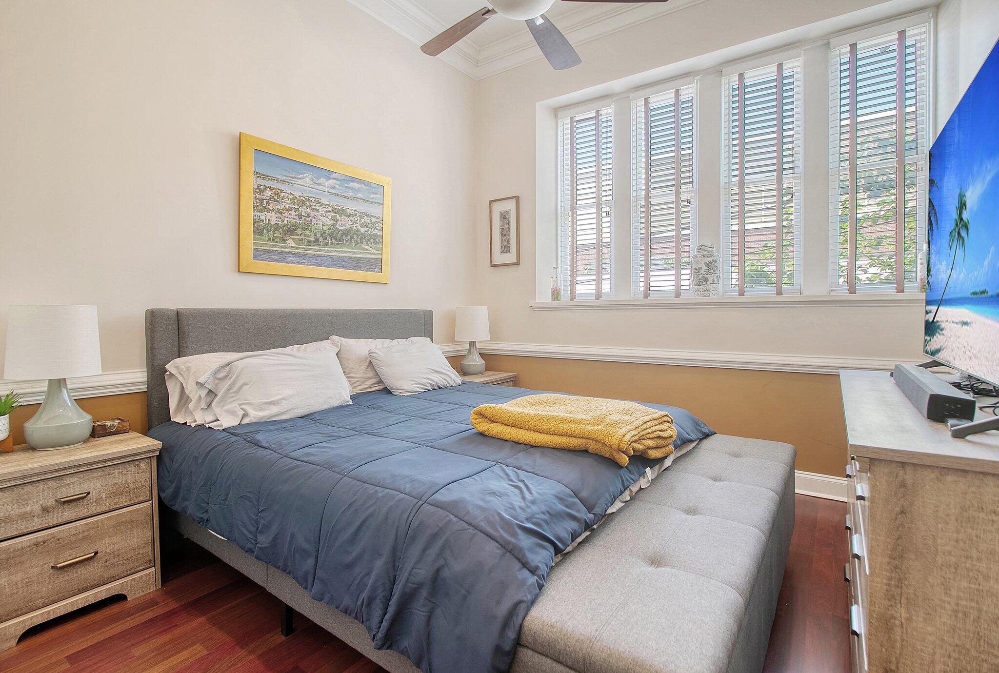 Bedroom with blue bedding, a gray upholstered bed and bench, and wide shuttered windows.