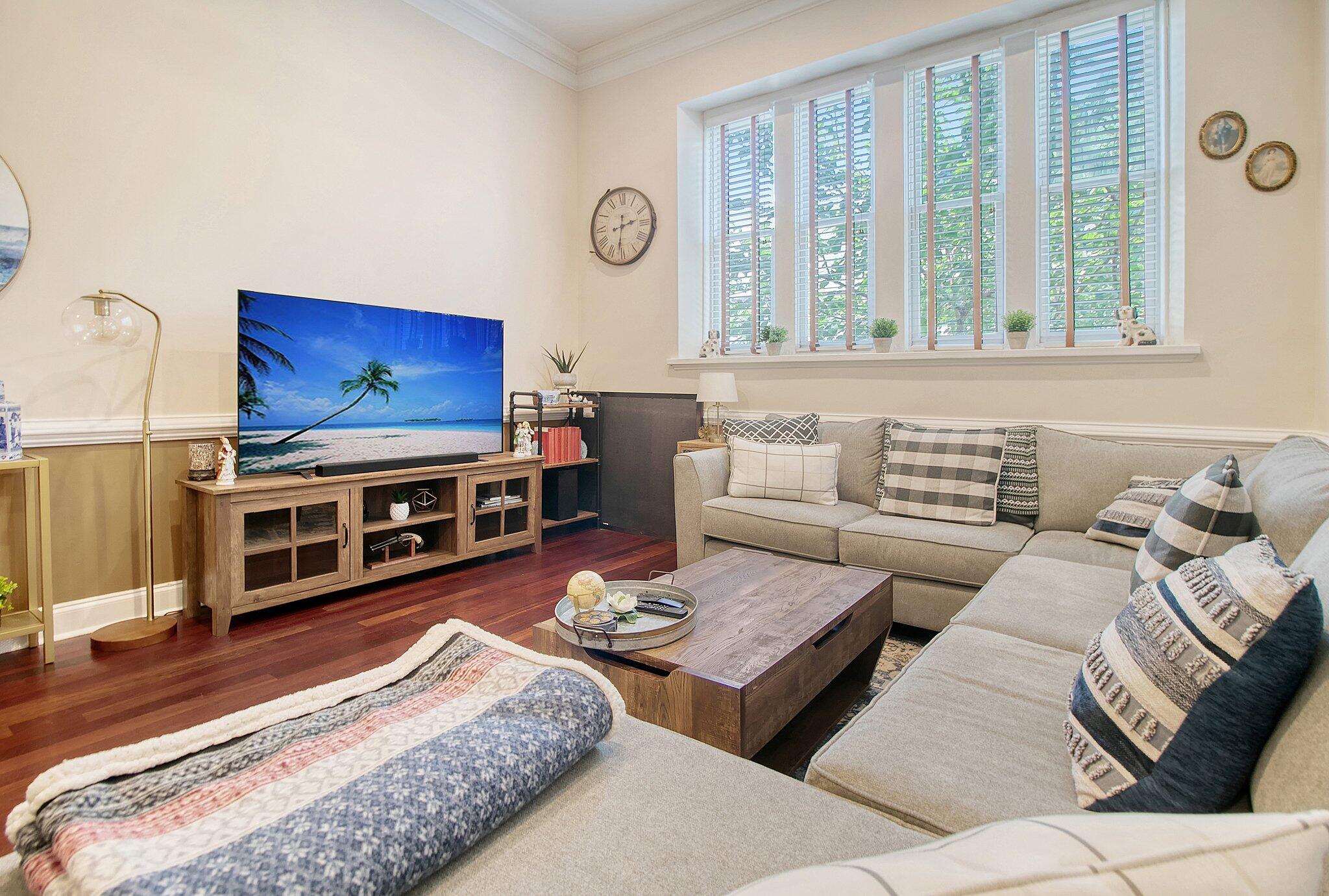 Furnished living room with a gray sectional, wooden coffee table, and television beneath wide shuttered windows.