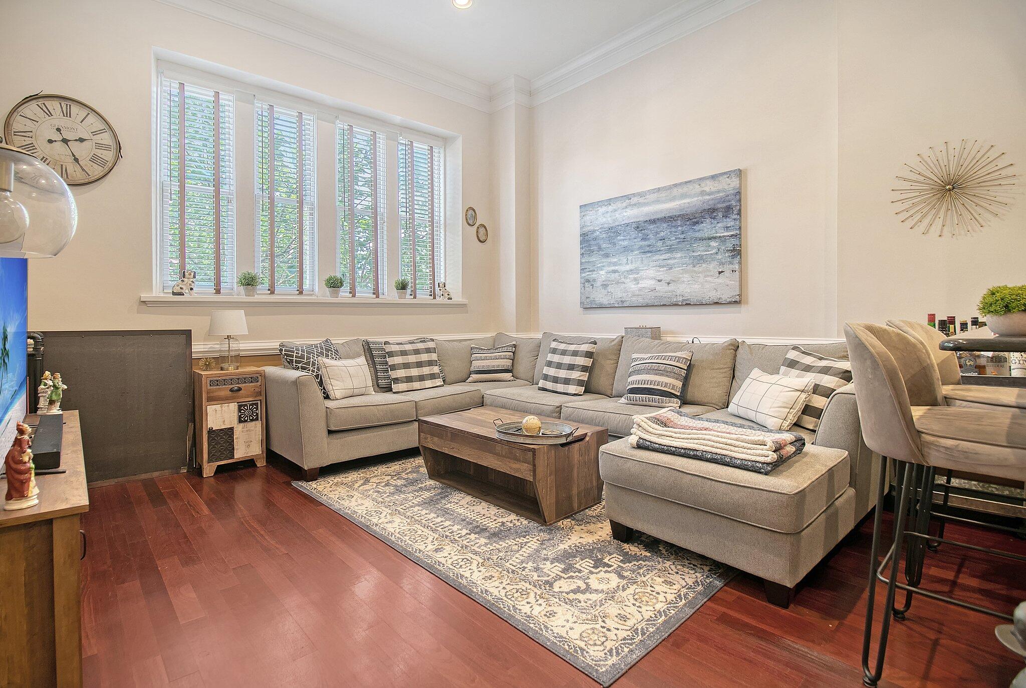 Living room with a gray sectional, patterned rug, dark hardwood floors, and shuttered windows.