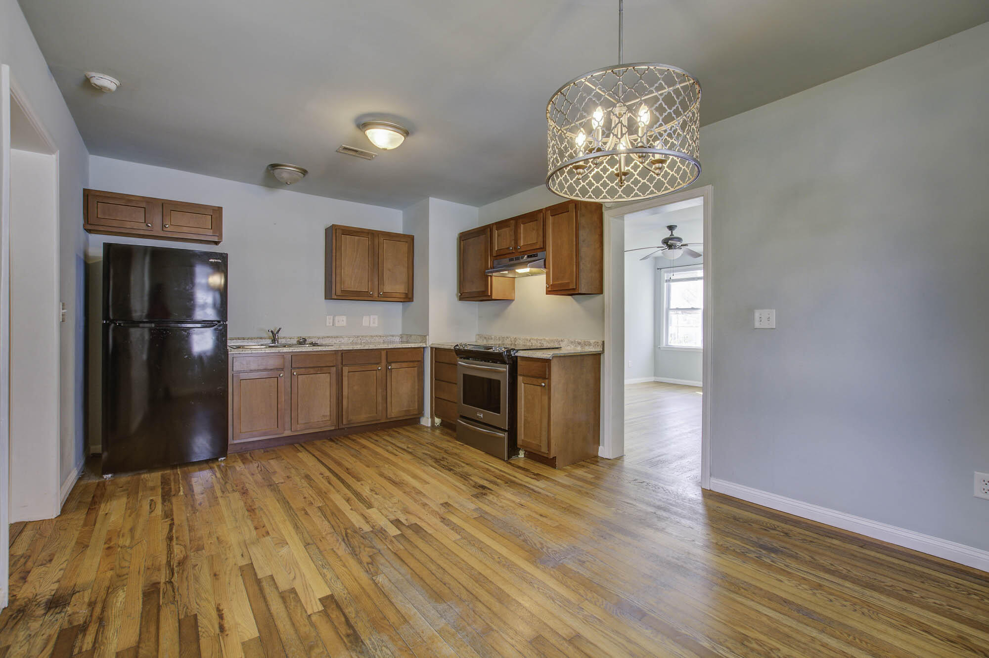 Vacant kitchen with reddish wood cabinets, black appliances, hardwood floors, and a patterned pendant light.