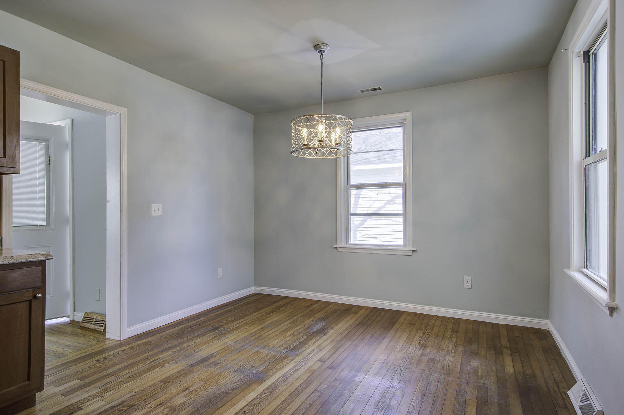 Empty dining room with hardwood floors, a decorative chandelier, and two windows.
