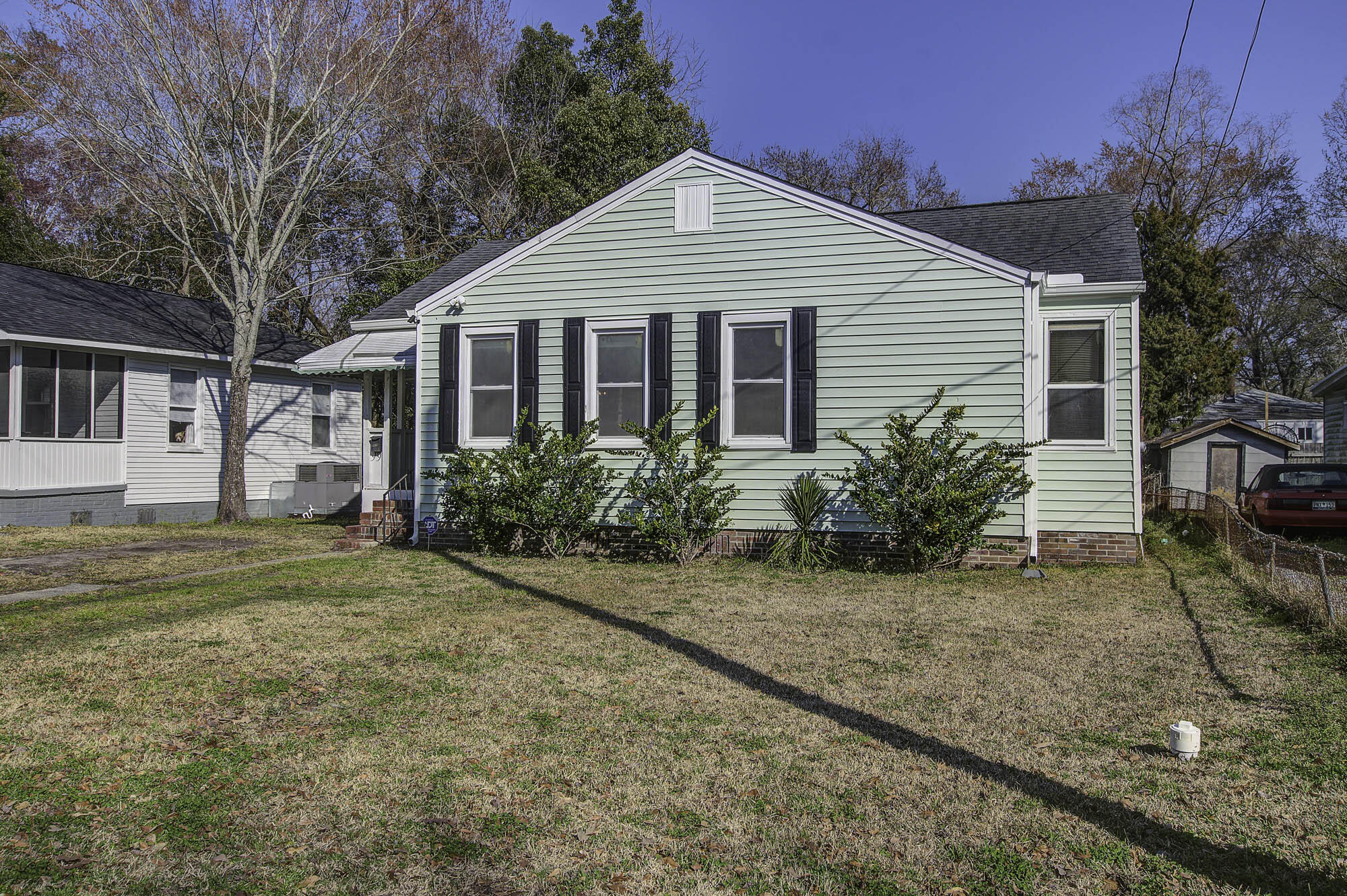 Sage-green single-story house with black shutters, front shrubs, and a broad winter lawn.