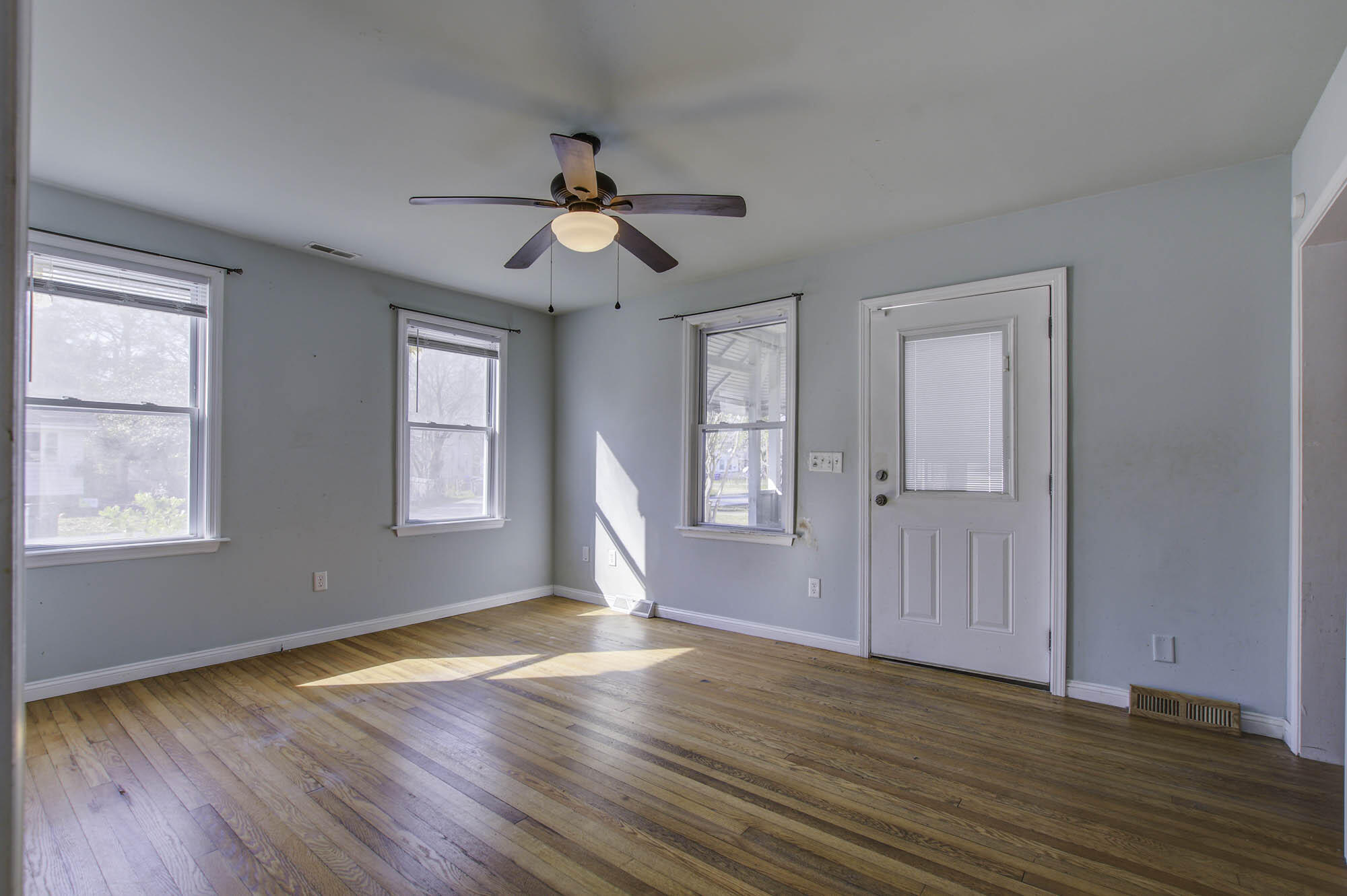 Vacant room with hardwood floors, pale blue walls, three windows, and a ceiling fan.