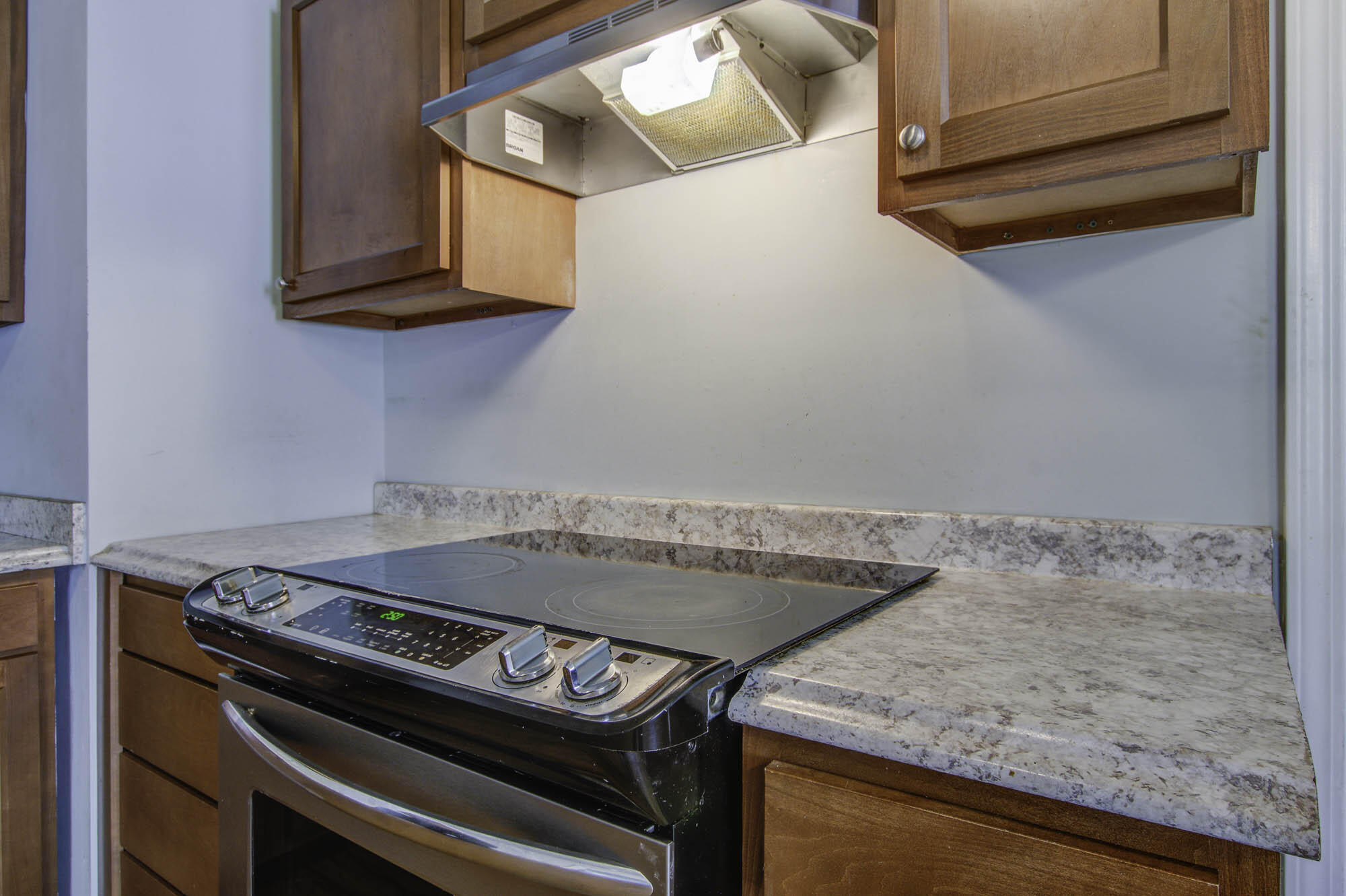 Kitchen range with glass cooktop, stainless steel oven, wood cabinets, and pale stone counters.