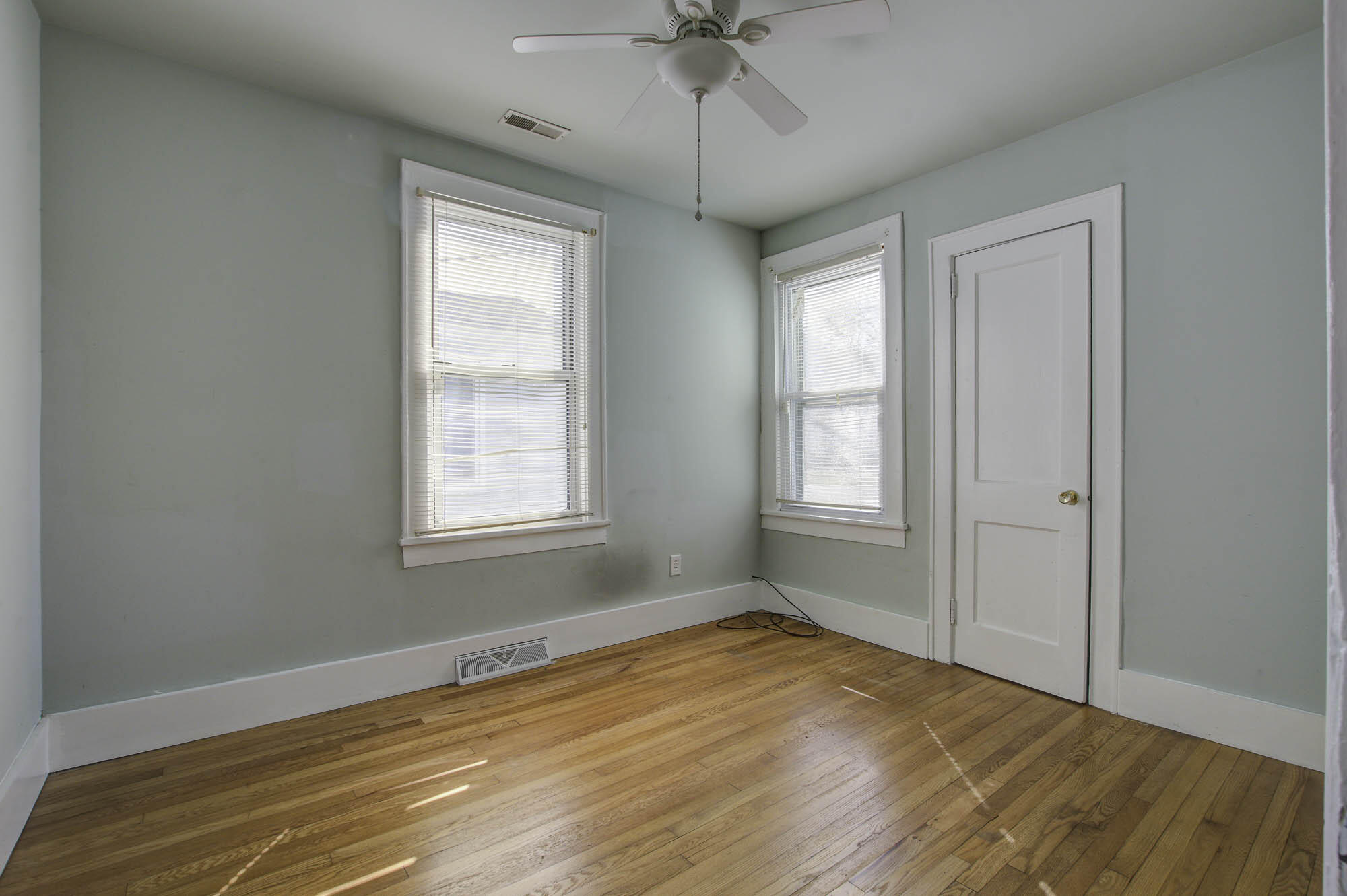 Empty room with hardwood floors, pale green walls, two blinded windows, and a ceiling fan.