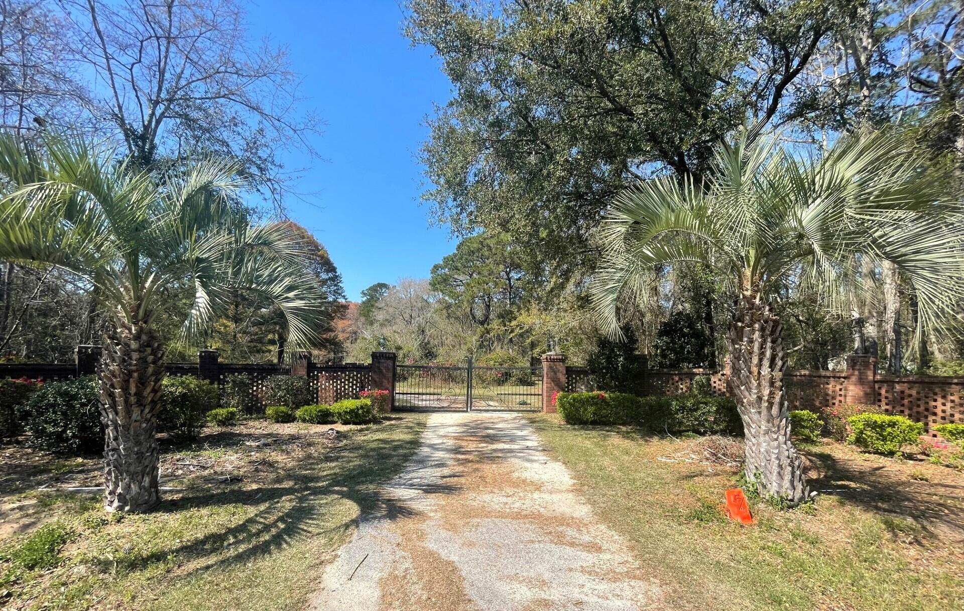 Gravel driveway and metal gate between brick walls, framed by palm trees.