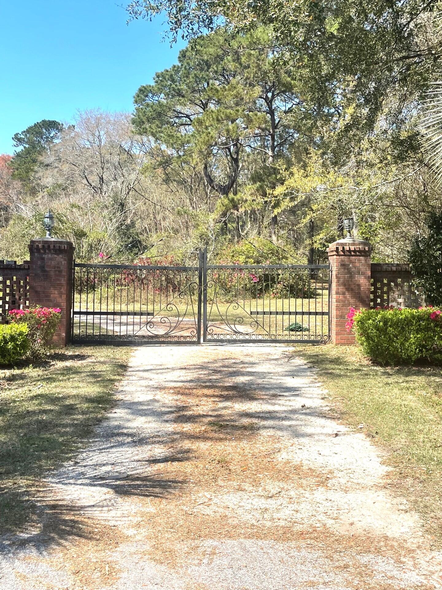Gravel driveway ending at an ornamental black gate between brick pillars, framed by flowering shrubs and trees.
