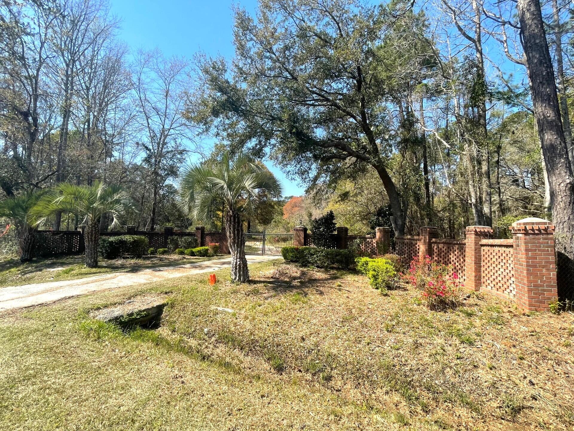 Gravel drive and metal gate beside a red brick lattice wall beneath mature trees and palms.