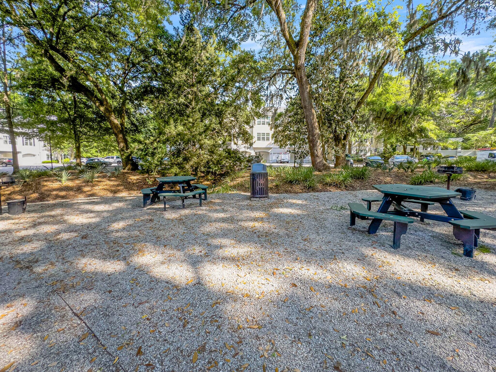 Shaded picnic area with two tables beneath mature trees, with apartment buildings in the background.