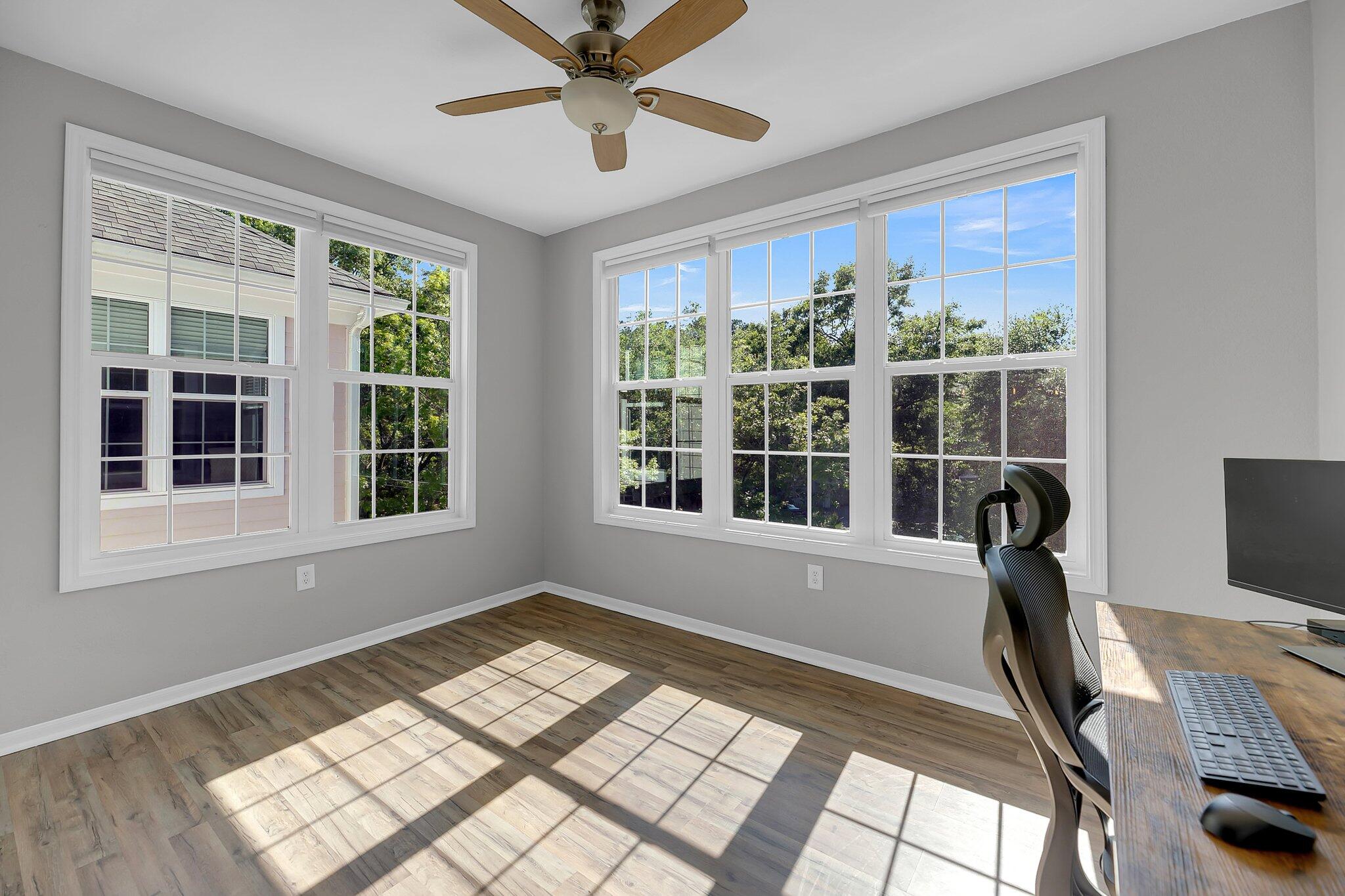 Home office with large sunlit windows, wood-look flooring, and a desk with an ergonomic chair.