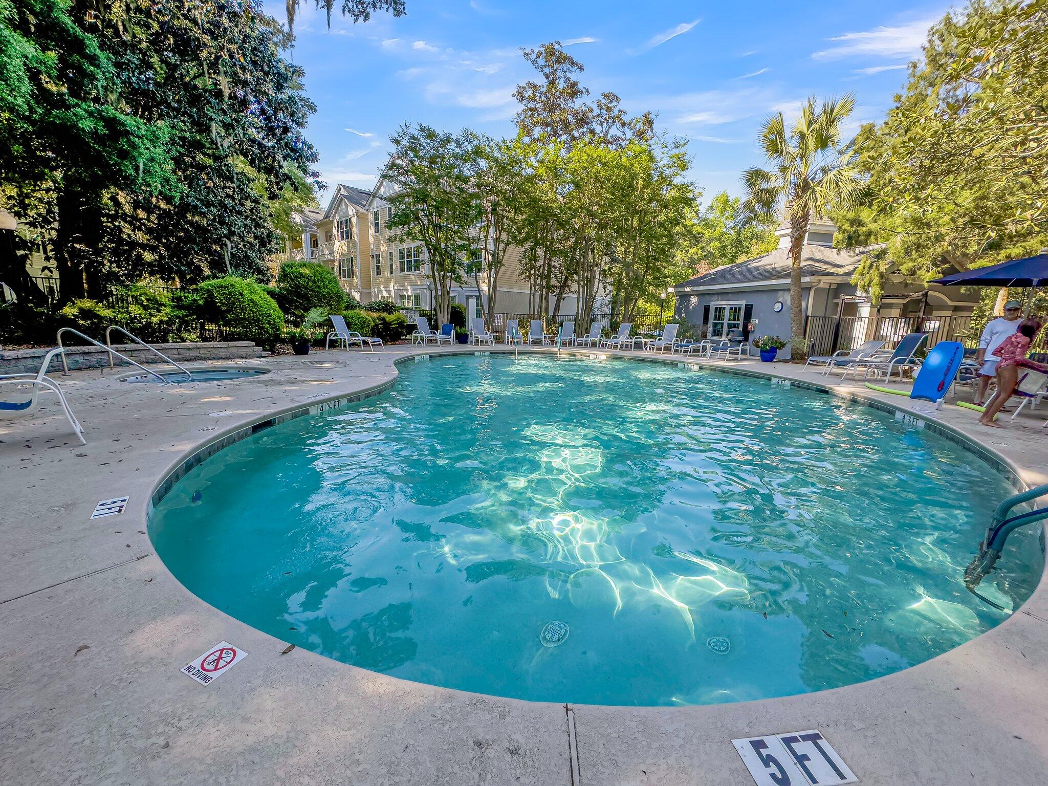 Community swimming pool with a curved deck, lounge chairs, and residential buildings among trees.