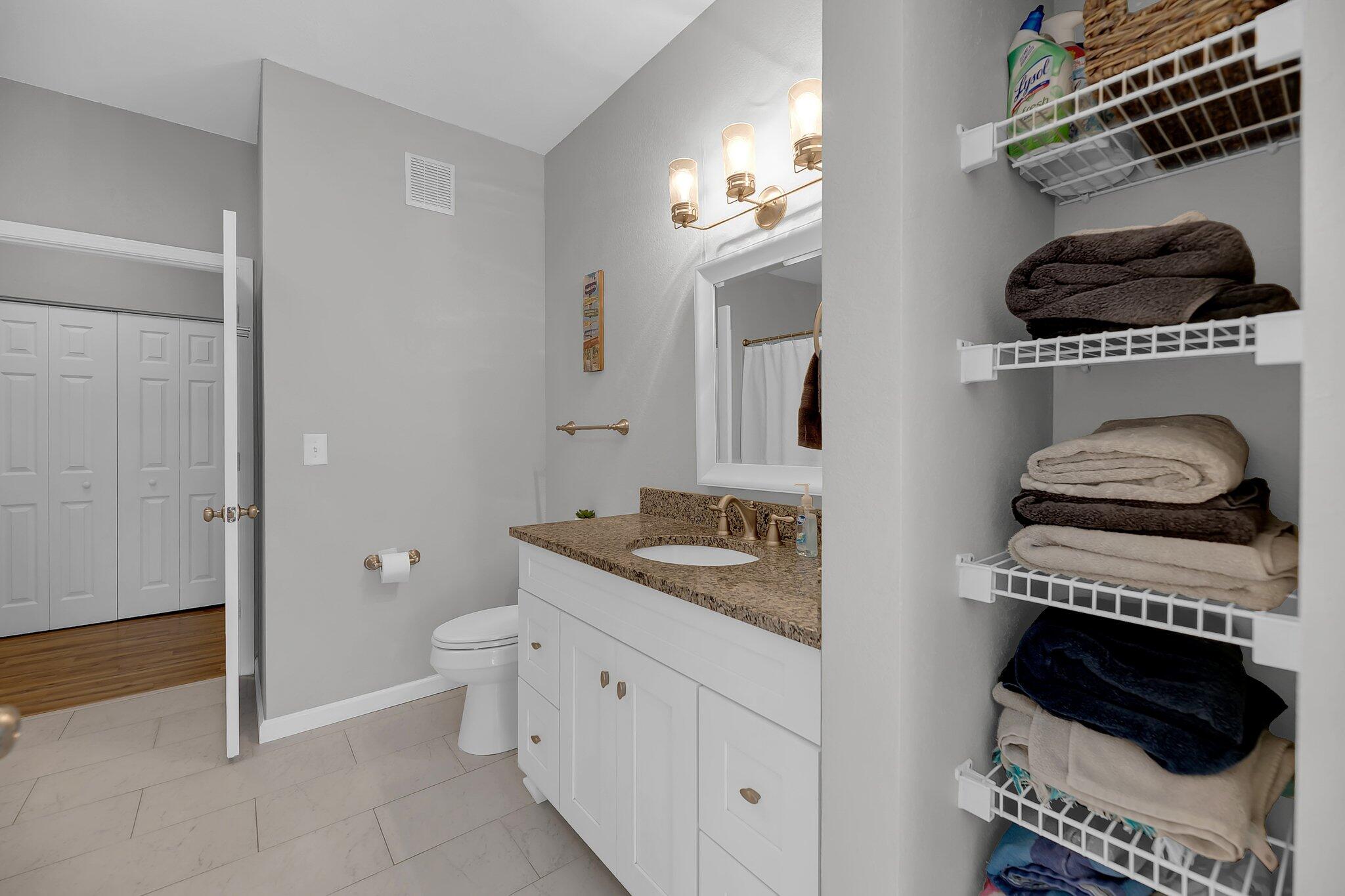 Bathroom with granite vanity, tiled floor, and open shelves stacked with folded towels.