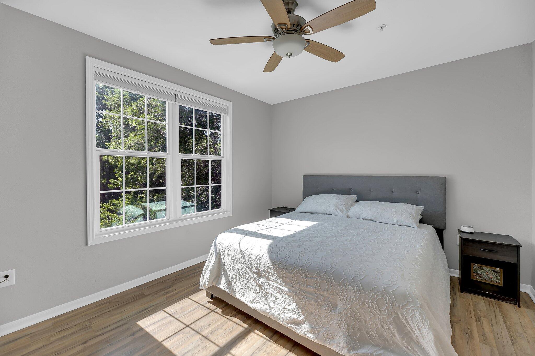 Bedroom with a large bed, gray headboard, wood-look flooring, and sunlit windows overlooking trees.