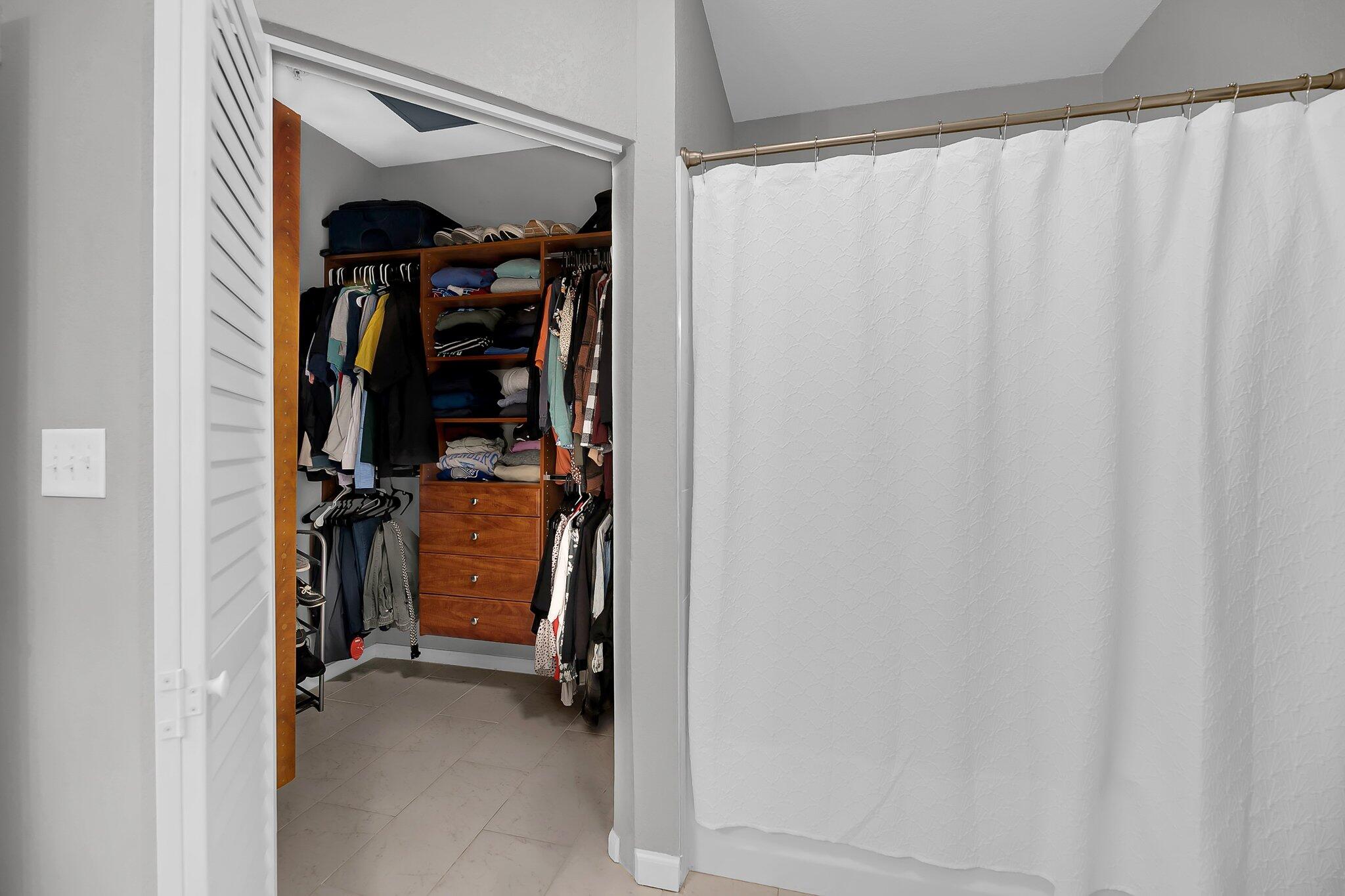 Walk-in closet with wooden drawers and clothing beside a bathroom shower curtain.