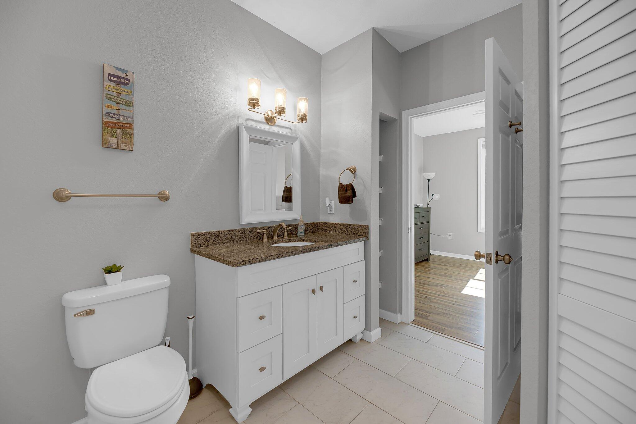 Bathroom with a white vanity, granite countertop, brass fixtures, and an open doorway to a wood-floored room.