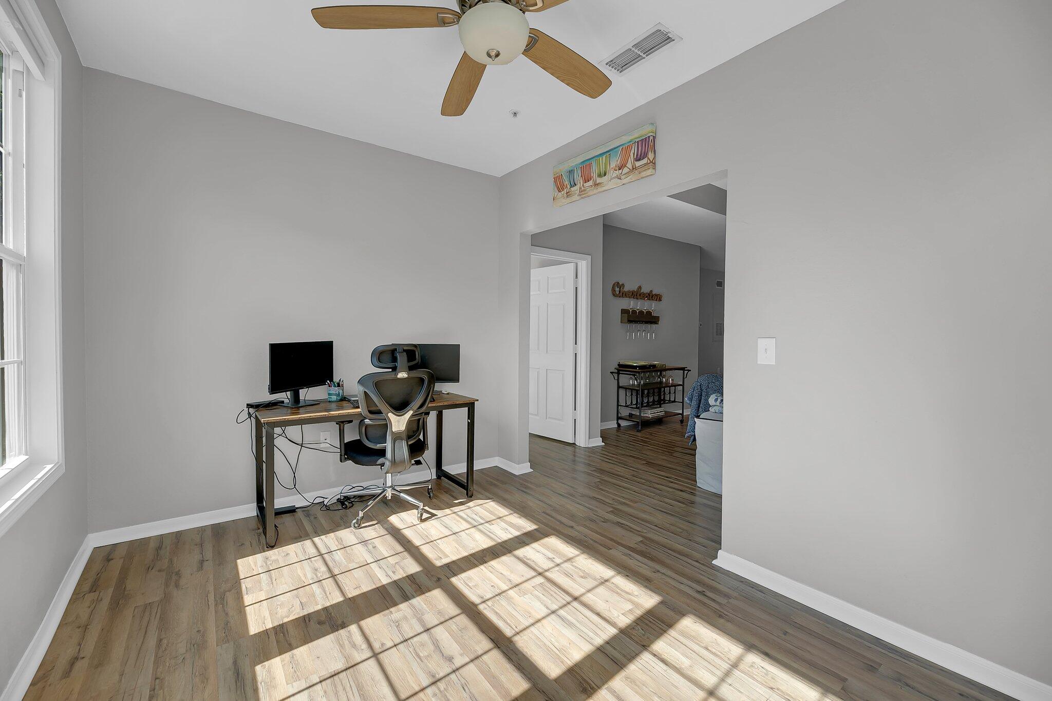 Home office with a desk and ergonomic chair, brightened by sunlight through tall windows.