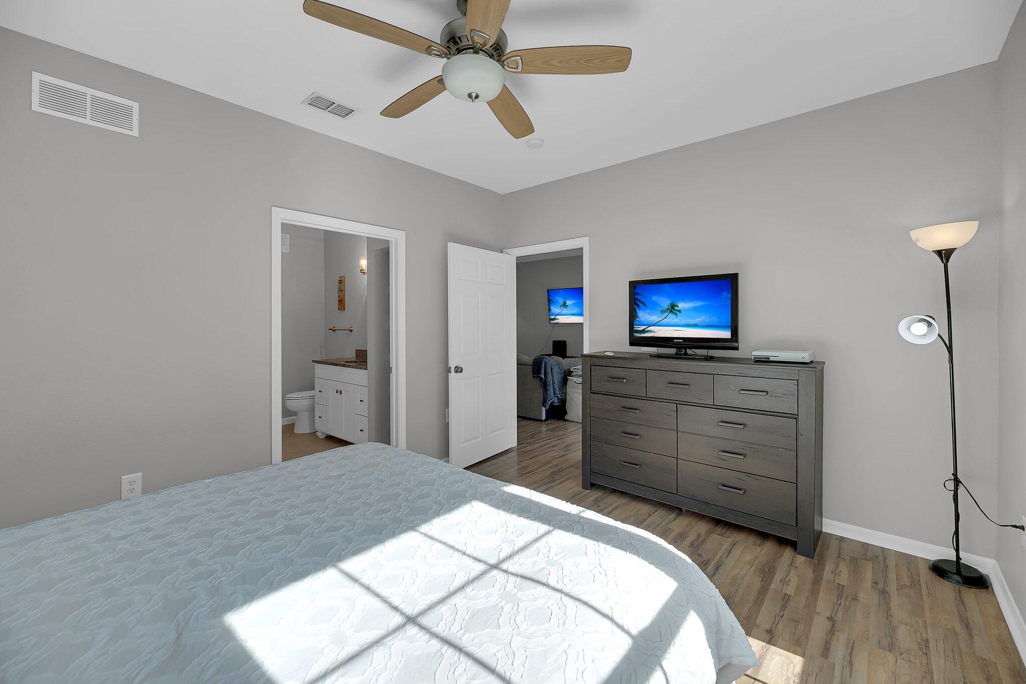 Bedroom with a white quilted bed, gray dresser and TV, and open doors to a bathroom and adjoining room.