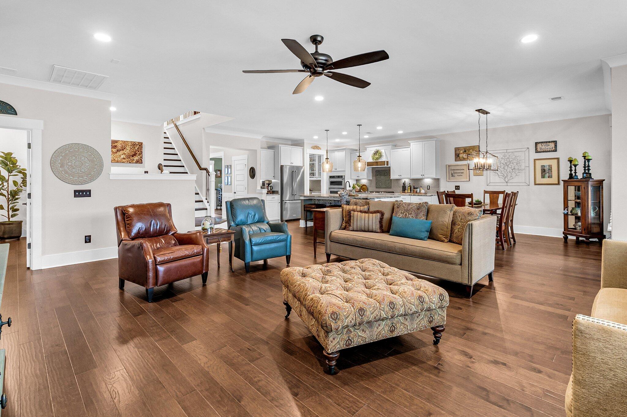 Open-plan living room with a beige sofa, patterned ottoman, hardwood floors, and the kitchen and dining area beyond.