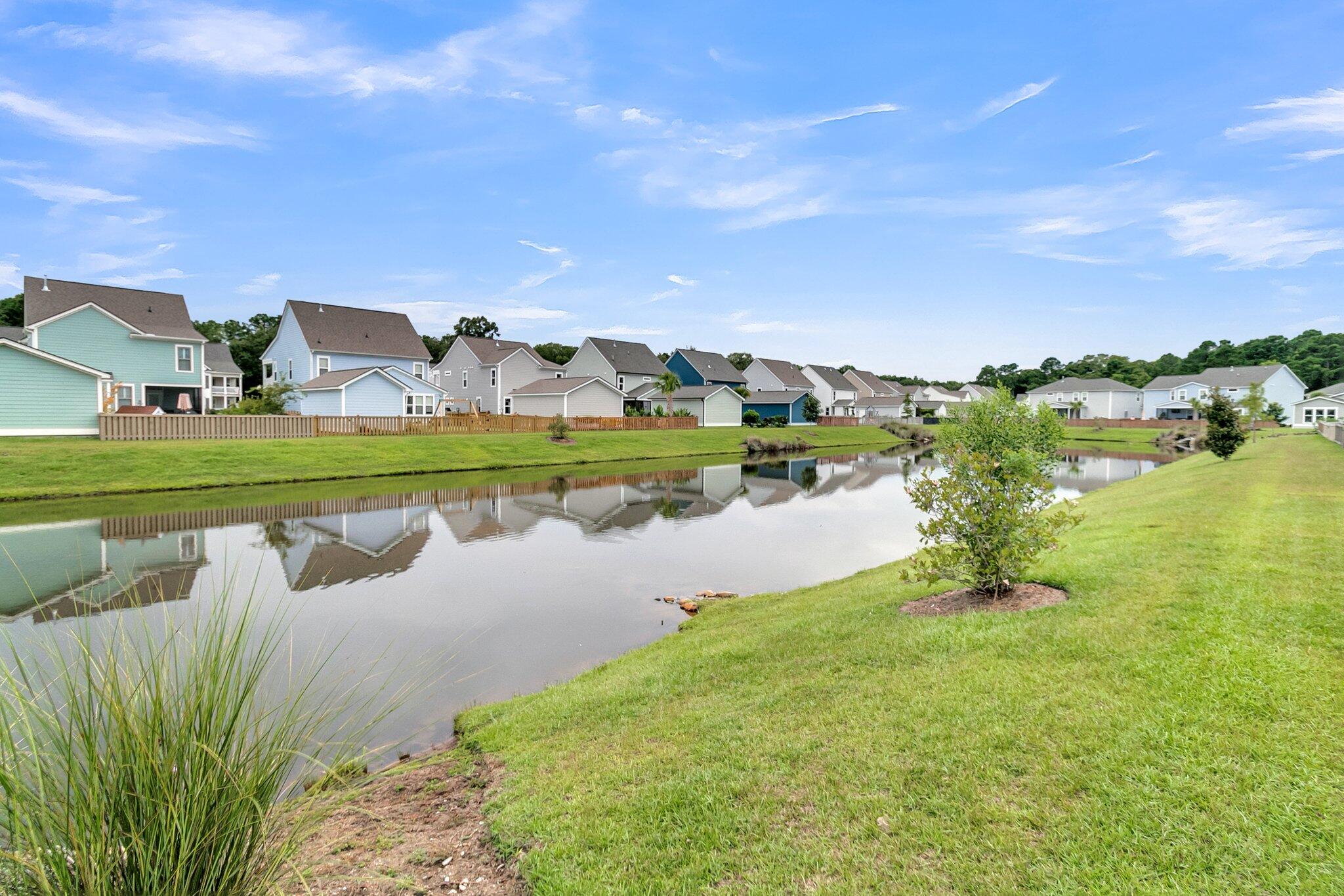 Neighborhood pond reflecting detached homes, with grassy banks and ornamental plants in the foreground.