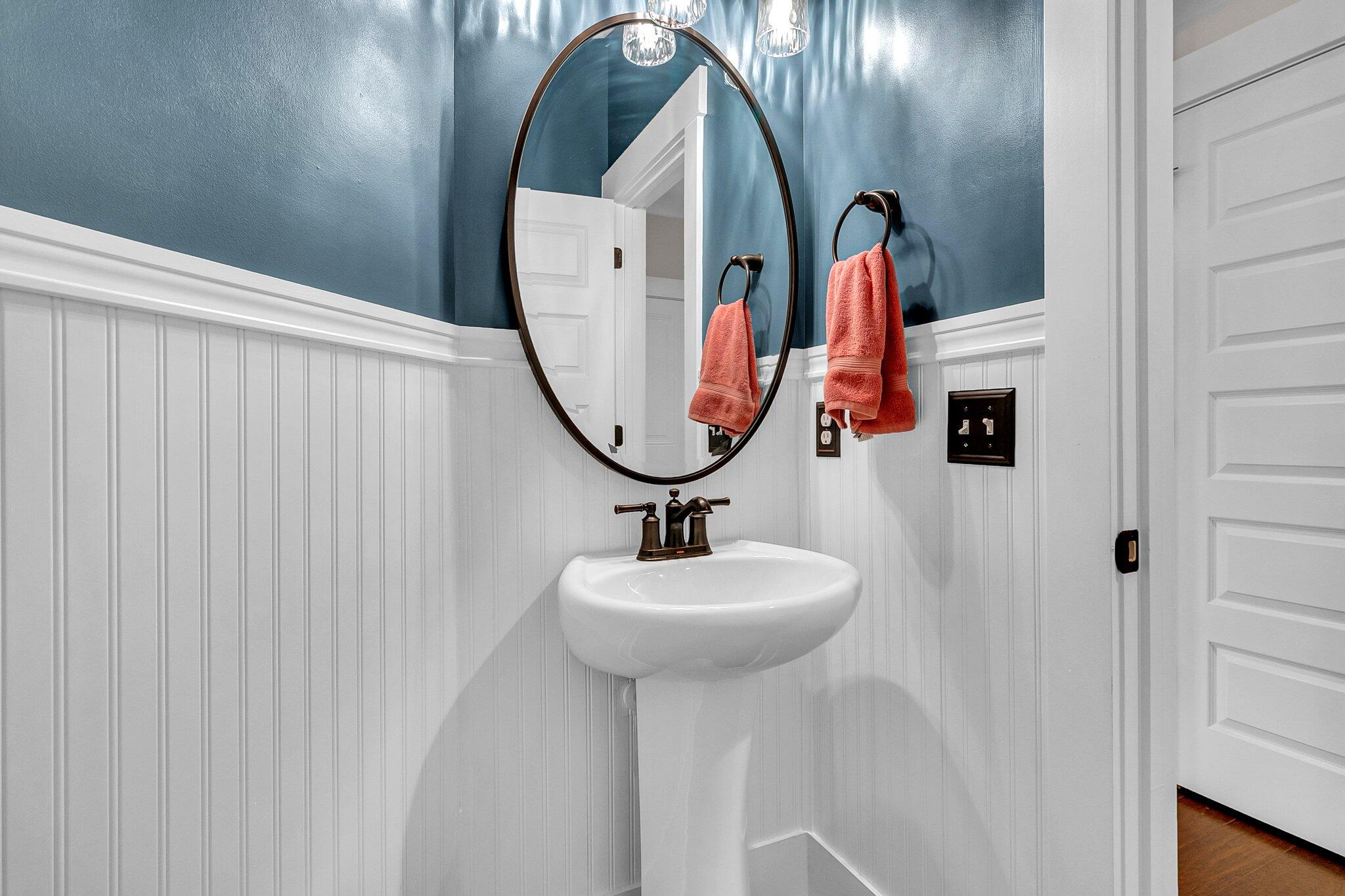 Bathroom with a pedestal sink, oval mirror, blue walls, white beadboard, and coral towels.