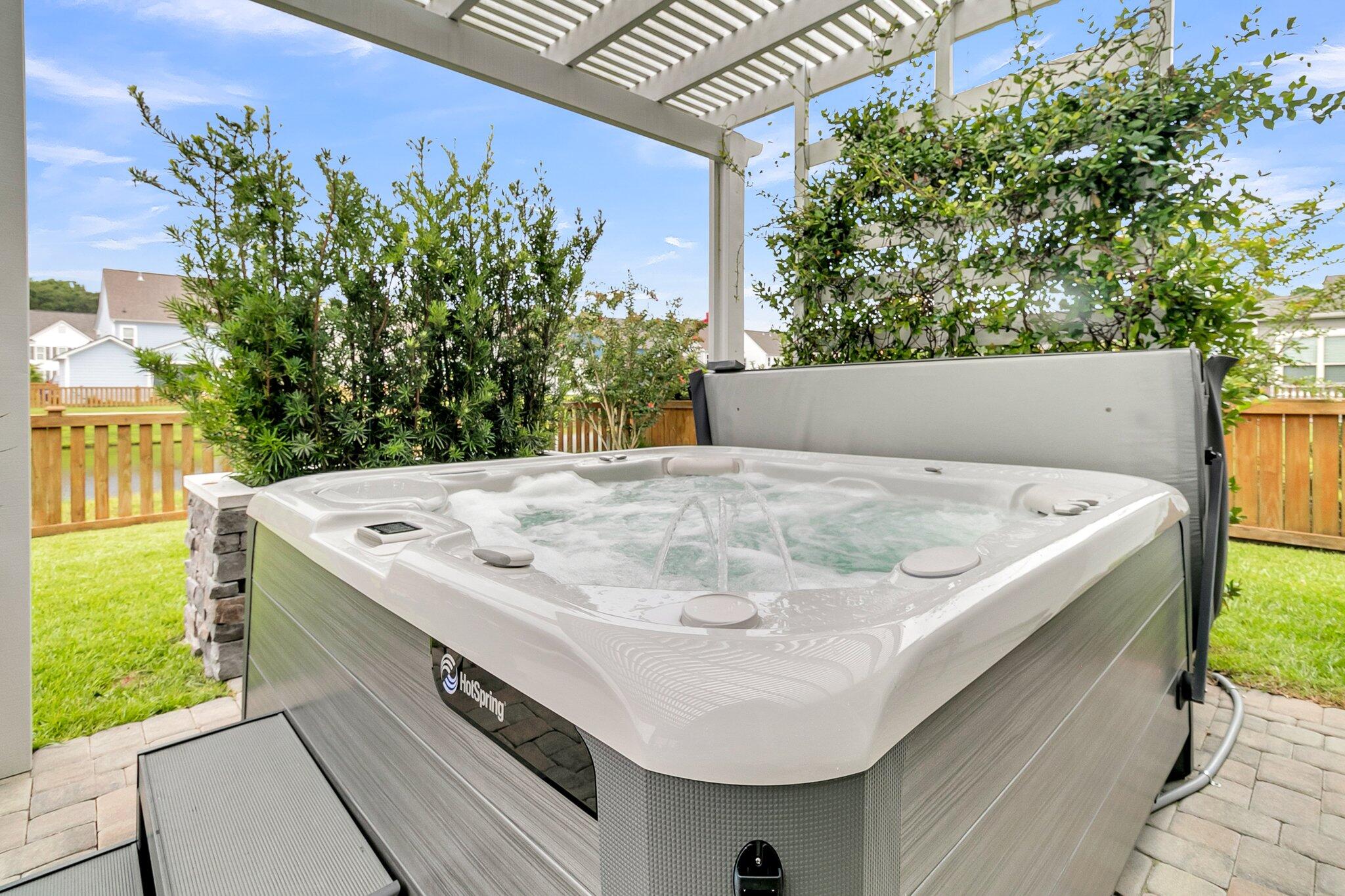 Hot tub bubbling beneath a white pergola on a paved patio, surrounded by shrubs and a fenced yard.