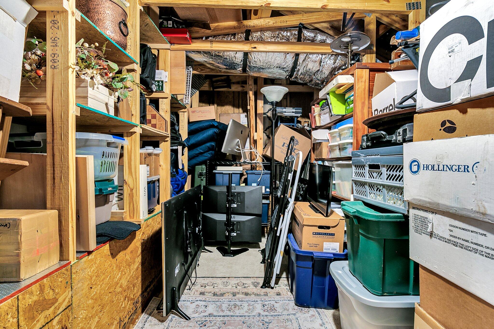 Crowded storage room with exposed framing, overhead ducts, shelves, boxes, and stacked plastic bins.