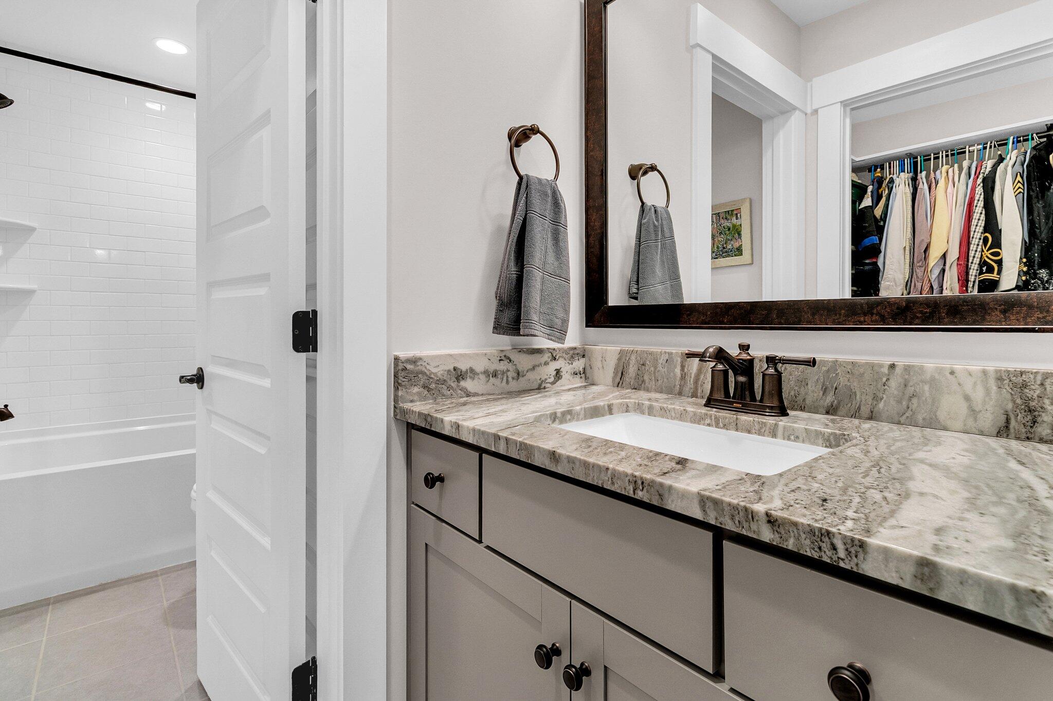 Bathroom with a gray vanity, veined stone counter, framed mirror, and doorway to a tiled tub.