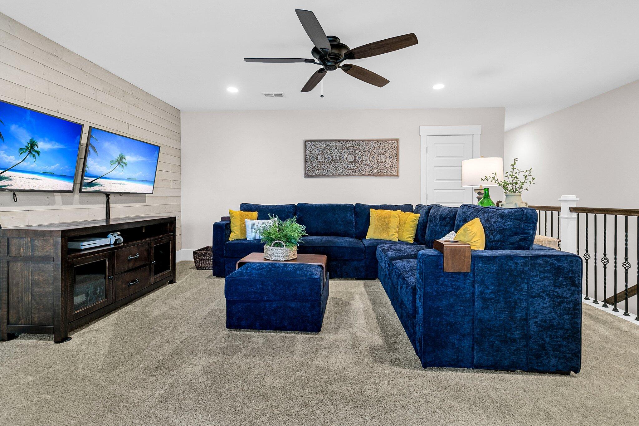 Living room with a navy sectional, yellow pillows, and two televisions on a shiplap wall.