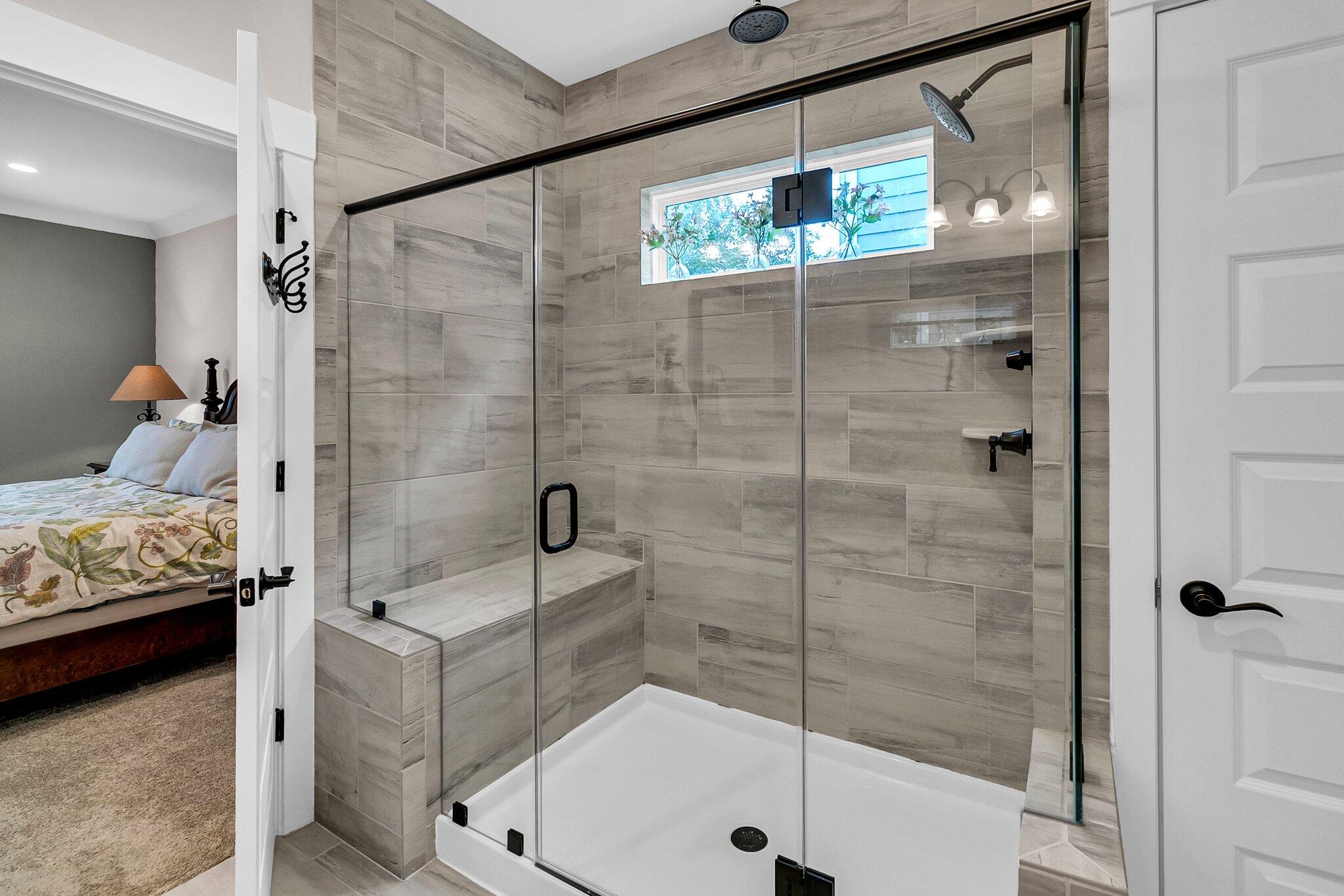 Walk-in shower with gray tile, glass doors, and a built-in bench beside a bedroom.