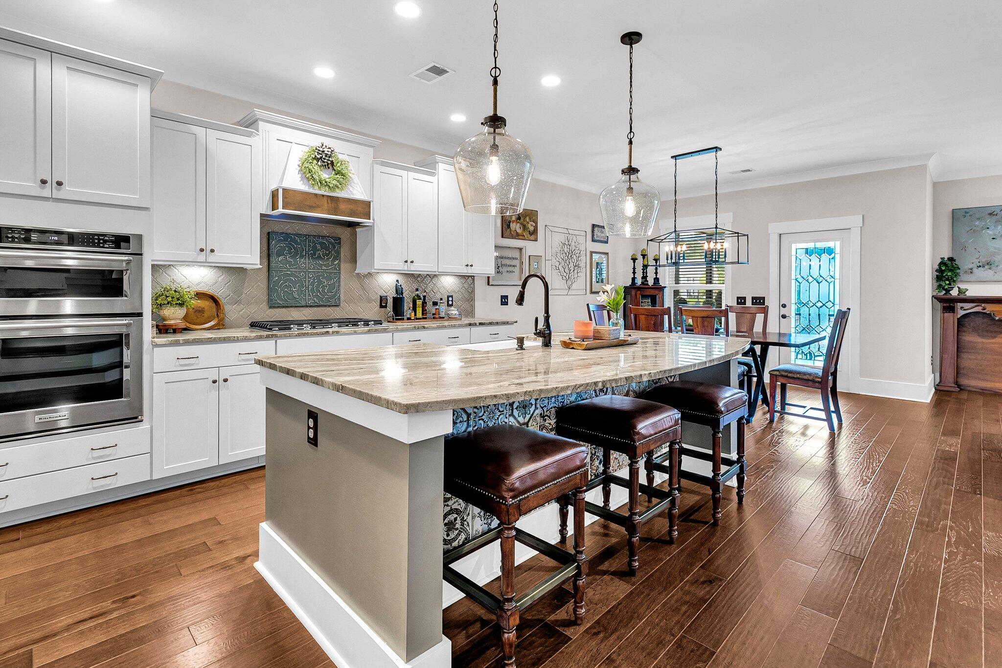 Kitchen with a large stone island, leather stools, white cabinets and patterned tile backsplash.