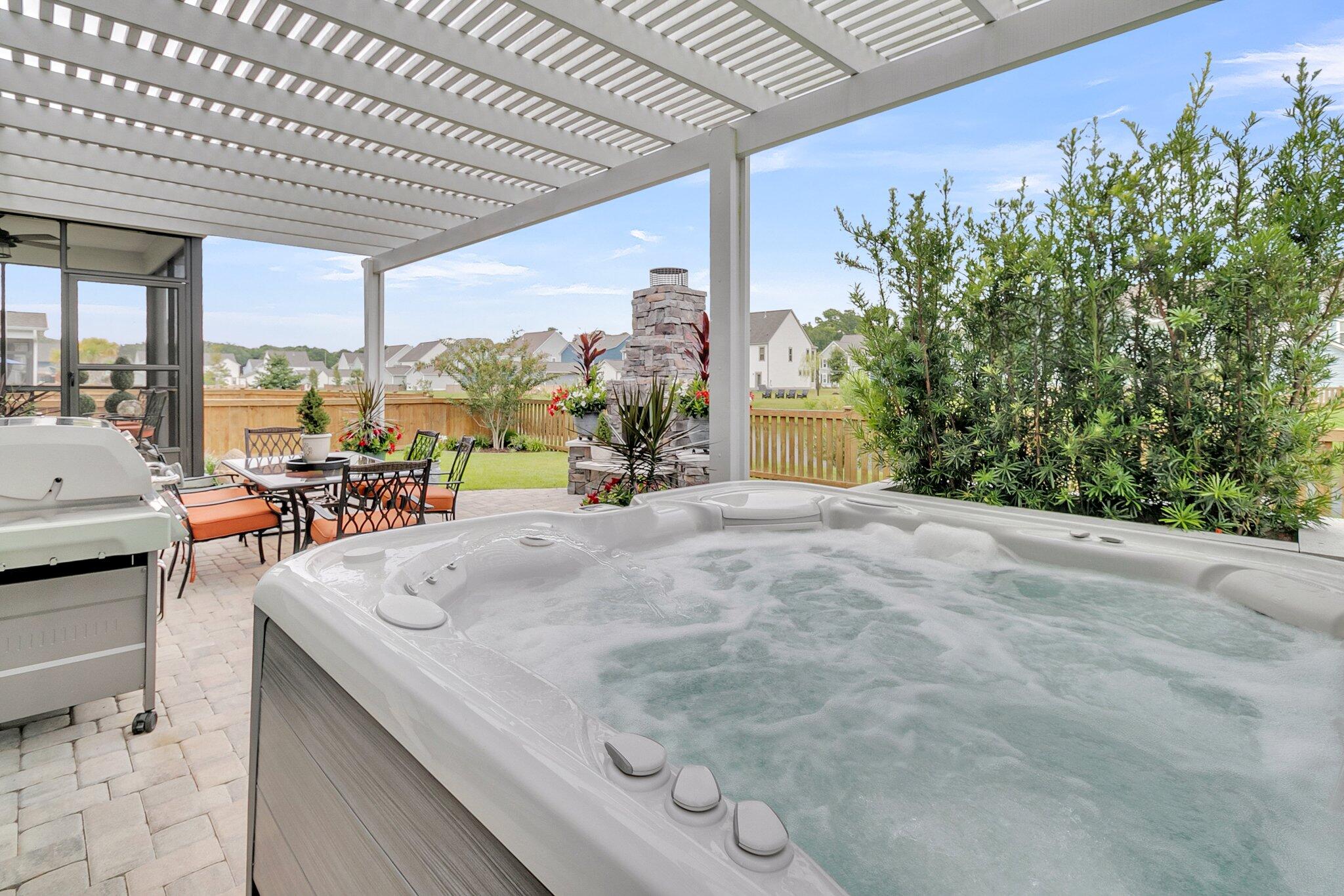 Backyard hot tub beneath a white pergola, beside outdoor dining furniture and a stone fireplace.
